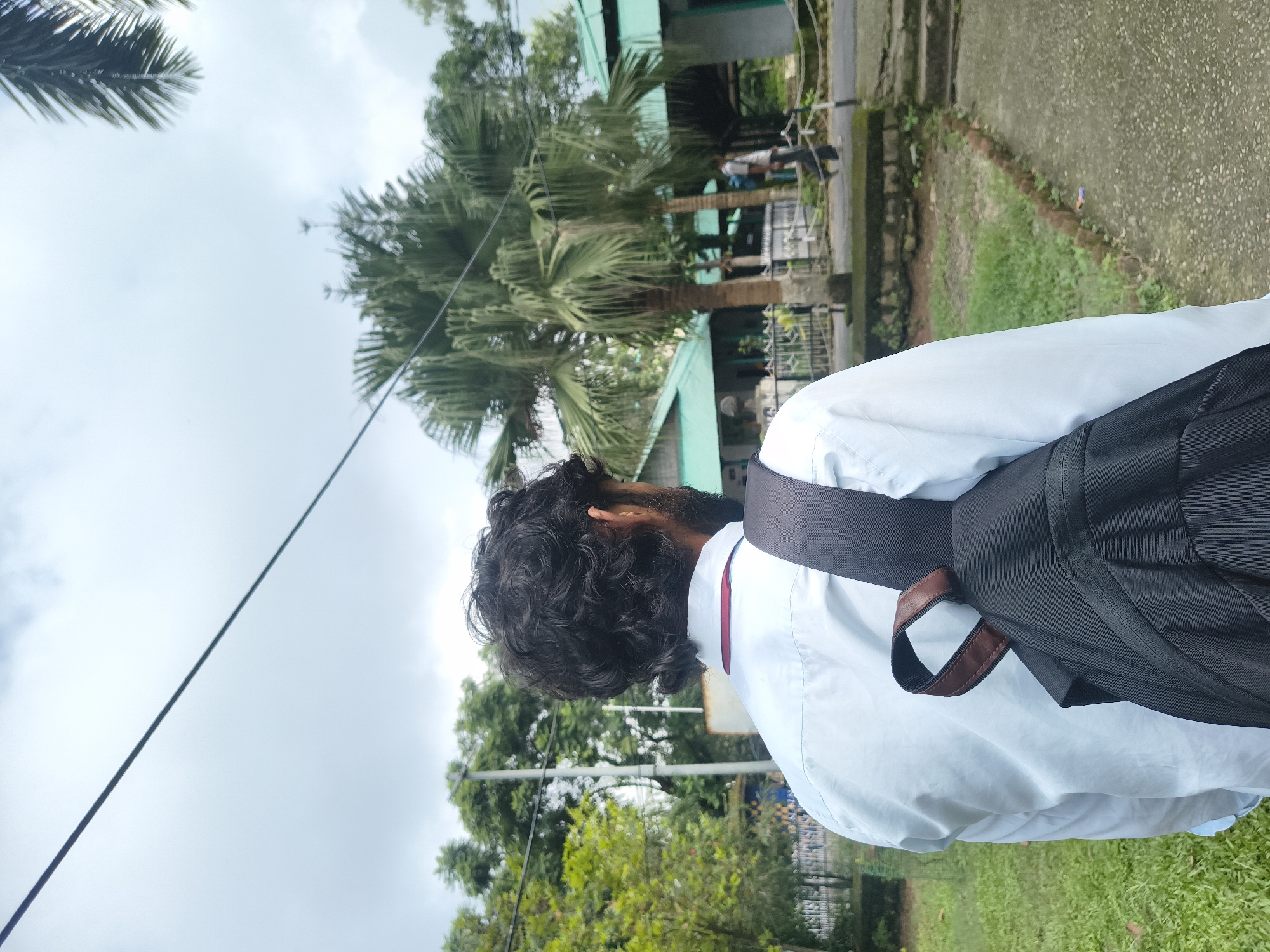 A person with curly dark hair and a beard, wearing a light blue shirt and a black backpack, stands with their back to the camera. They are looking towards a building with a green roof, surrounded by lush green trees and palm trees. The sky is overcast. In the background, another person is walking away from the camera on a path. The scene appears to be outdoors, possibly in a park or a campus area. The location is identified as Dharamshala, India.