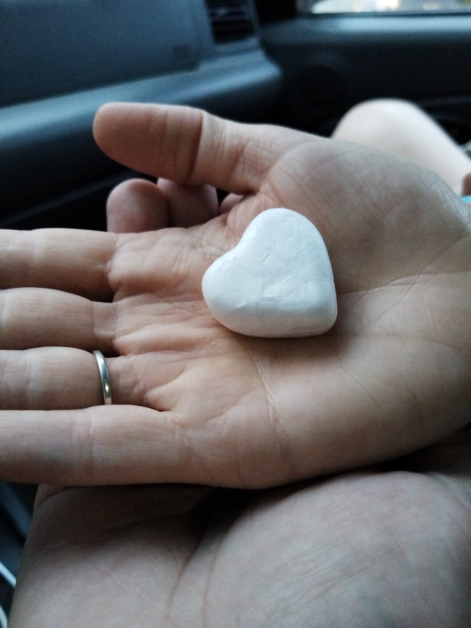 A person's hand is holding a small, white, heart-shaped object. The hand has a silver ring on one of the fingers. The background appears to be the interior of a car, with a portion of the dashboard visible. There is no information to indicate that this picture was taken in the city of Chaikovsky, Russia.