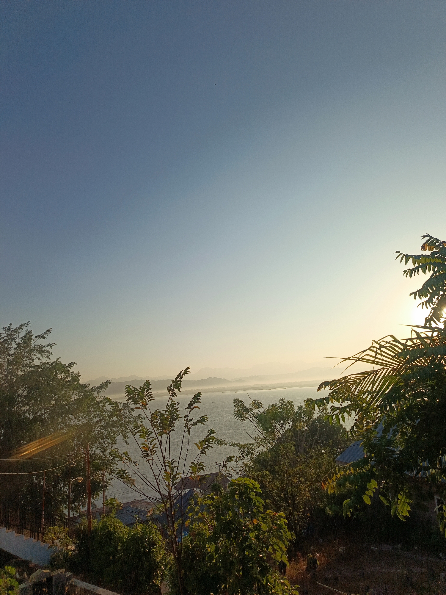 The image depicts a serene vista overlooking a body of water, likely a bay or lake, with mountains in the distance, at sunrise or sunset. The sky transitions from a clear blue at the top to a warm, hazy yellow near the horizon, illuminated by the sun peeking through foliage. In the foreground, lush green trees and branches frame the scene, with some foliage silhouetted against the bright sky. A hint of structures, possibly rooftops and utility poles, can be discerned beneath the trees on the left side of the image, suggesting a residential or semi-urban area. The overall atmosphere is tranquil and natural, bathed in the soft light of dawn or dusk. The location is identified as the City of Gorontalo, Indonesia, a region known for its coastal beauty and surrounding mountainous terrain.