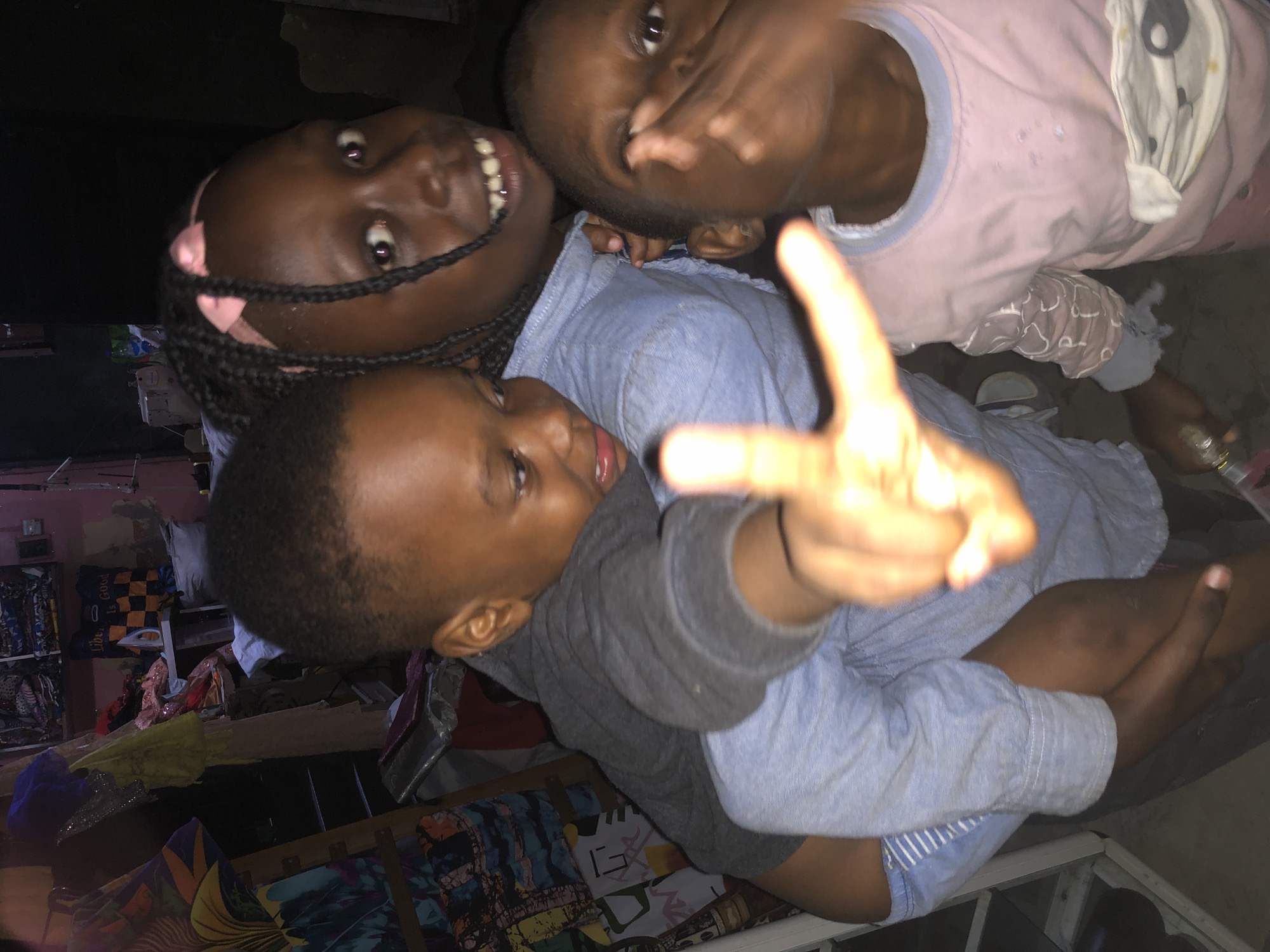 The image depicts a dimly lit indoor environment, likely a room in Ado, Nigeria, characterized by shelves filled with colorful textiles and clothing items, suggestive of a tailor's shop or a market stall. Three children are prominently featured. In the foreground, a young boy is being carried on the back of another child, possibly an older sister. He is looking downwards and to his left, with his right hand extended forward, making a "peace" sign. The child carrying him is a girl with braided hair and a pink headband, looking directly at the camera with a wide smile. To the right of these two, a third child, also a young boy, is partially visible, looking towards the camera and appearing to be making a gesture with his hand. The overall impression is one of a candid, spontaneous moment captured amidst a busy, domestic setting.