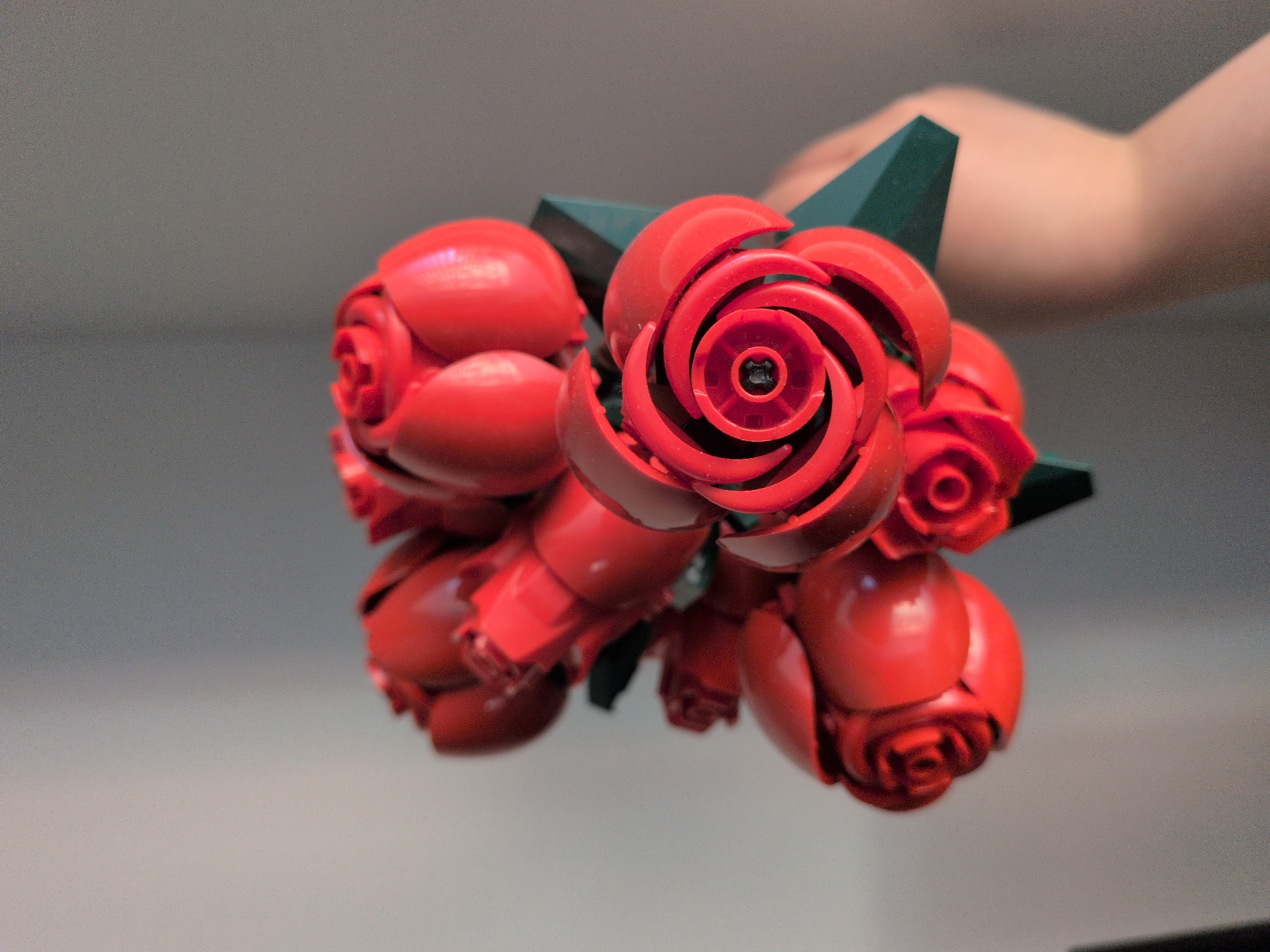 A high-angle, close-up photograph displays a bouquet of artificial red roses held by a person's hand in the city of Unknown, Unknown. The roses are constructed from bright red plastic, with distinct, rigid petals arranged in spiraling patterns, characteristic of building toy elements. The most prominent rose in the foreground features a dark circular component within its center. Multiple roses and buds are clustered together, with dark green, angular plastic pieces forming the base of the bouquet where it is held. A light-skinned right hand, visible from the wrist to the lower forearm, supports the bouquet. The background is a plain, light grey wall, softly lit, providing no additional environmental context or identifying features.