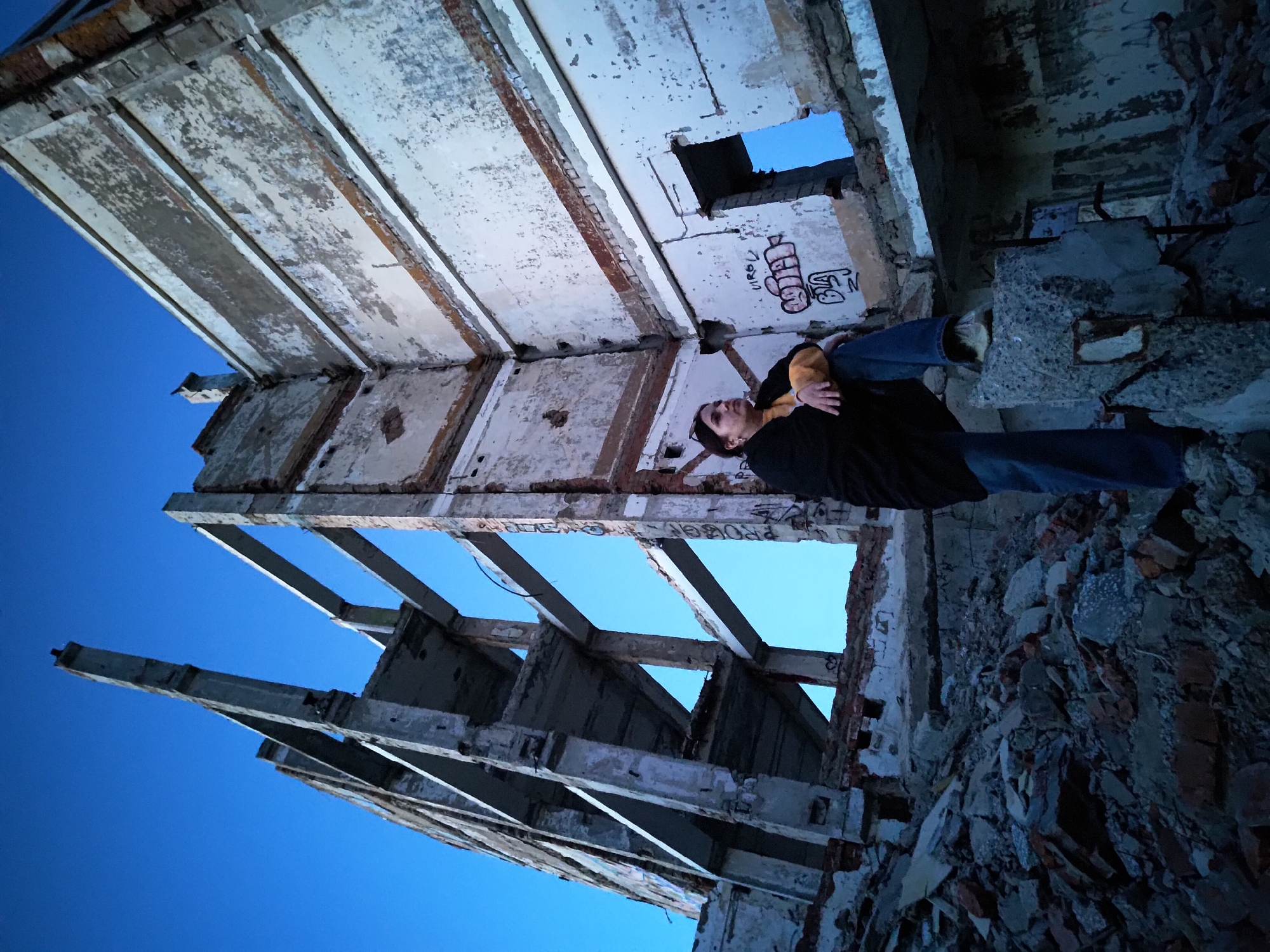 The photograph, taken from a low angle, depicts a person standing amidst the ruins of a derelict building. The sky is visible through the building's decaying skeleton.
The person is wearing jeans, a yellow shirt, and a black jacket. They are standing on a pile of rubble, composed of broken bricks, concrete, and other debris.
The building's structure is severely damaged, with parts of the walls and roof missing, exposing the open sky. Patches of peeling white paint and graffiti adorn the remaining walls. Based on the graffiti tags, this image was taken in Марковский, Russia.
