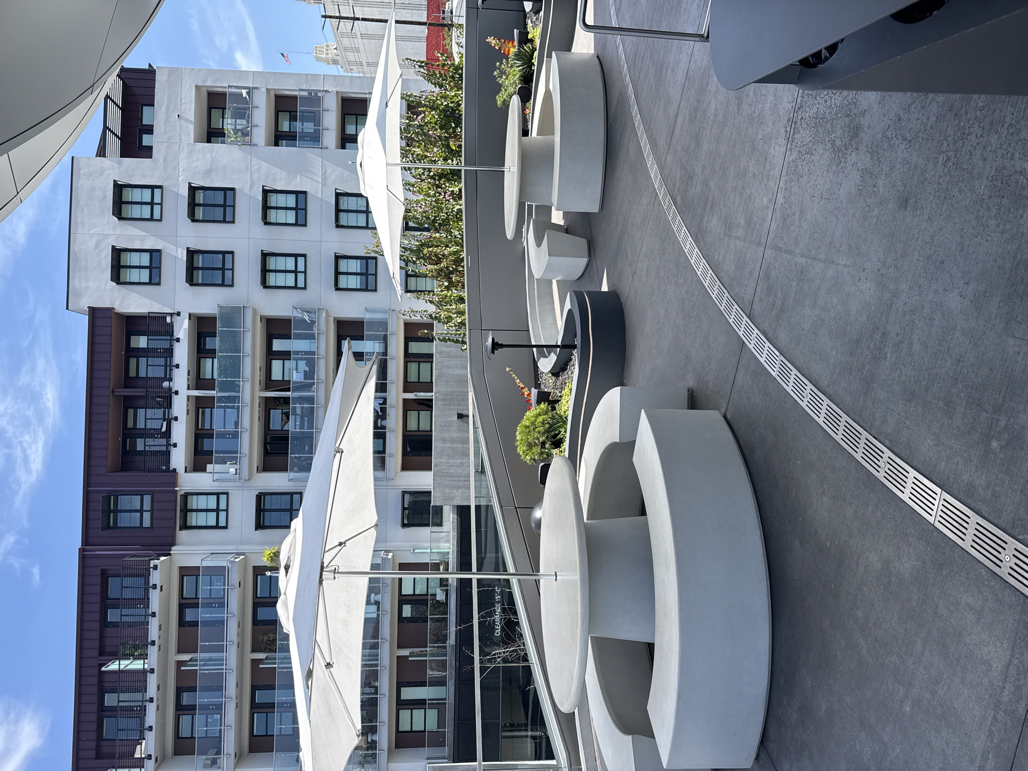 This outdoor shot shows a series of modern, curved concrete benches and tables situated on a gray, textured surface, likely a patio or rooftop area. The benches are arranged in semi-circular shapes, with smaller, round stools accompanying the tables. Some of the tables are paired with sun umbrellas. A linear drain runs along the patio floor.
In the background, a large, multi-story apartment building is visible, featuring a facade with repetitive window patterns, balconies, and a mix of white and brown architectural elements. The building's style is contemporary, and the presence of the building, combined with the upscale patio furniture, indicates an urban setting. The sky is blue with some clouds.
While the prompt indicates the location as West Hollywood, United States of America, this information is not visually verifiable from the image alone.