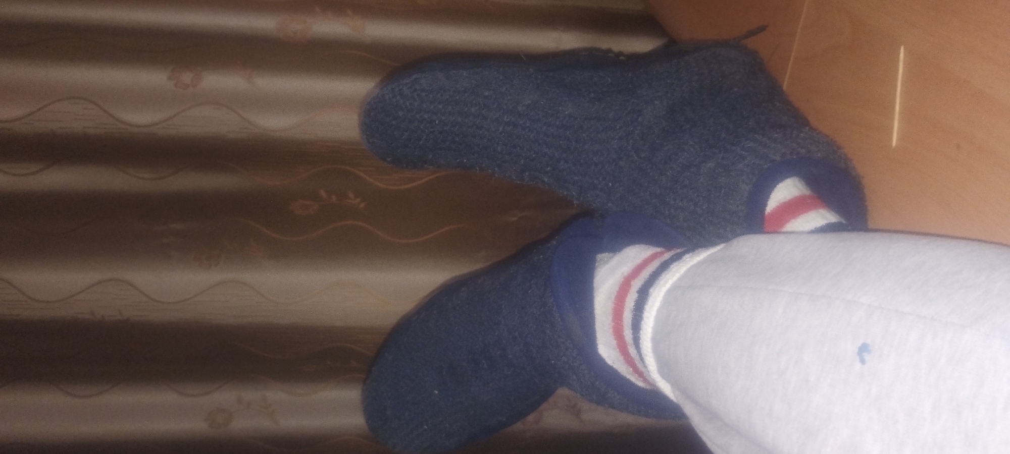 The media file shows a close-up view of a person's feet wearing dark blue knitted slippers over white socks with red and blue stripes. The slippers are covering the feet and ankles, and the socks are visible peeking out from the top of the slippers. The person's legs are also visible, clad in light grey sweatpants.
The scene appears to be indoors, with a brown, textured surface in the background, possibly a curtain or a fabric-covered wall, which has a subtle wavy pattern. To the right of the feet, a light brown wooden surface is visible, likely a piece of furniture like a bed or a table. A single, thin, light-colored stick, possibly a toothpick or a skewer, rests on the wooden surface.
The lighting suggests it might be daytime, though the exact time is not discernible. There are no clear indications of the weather or the specific location, other than it being an indoor environment. The image is focused on the feet and lower legs, and no emotions are conveyed. There is no visible text in the image. The overall impression is a casual, domestic setting.