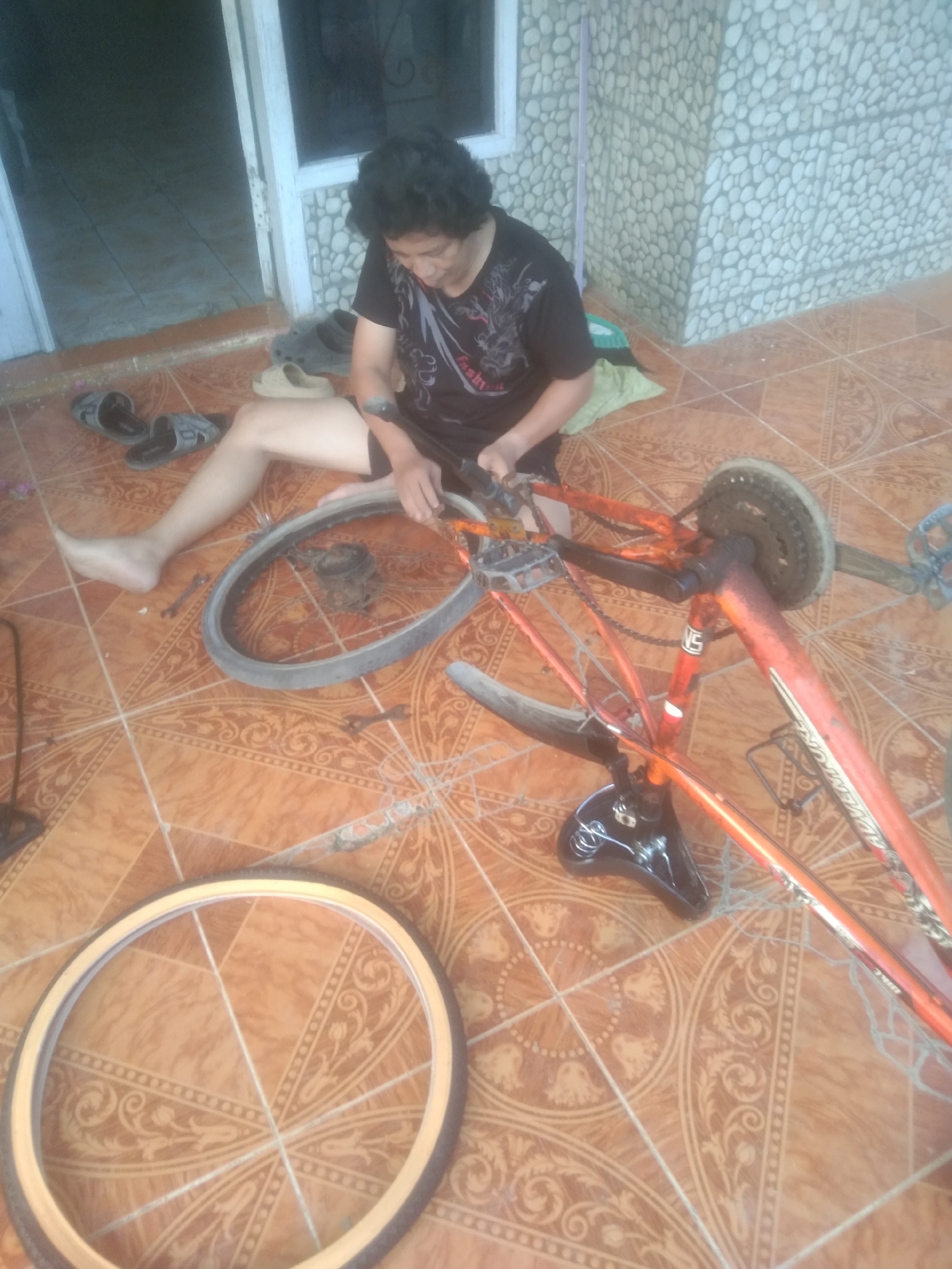 A person with dark, curly hair is working on an orange bicycle on a tiled patio in Subang, Indonesia. The person is wearing a black t-shirt with a graphic design and black shorts. They are seated with their legs extended and are holding tools. The bicycle is partially disassembled, with the front wheel removed and resting on the ground in the foreground. The rear wheel is still attached to the frame. Other bicycle parts, including the chain and gears, are visible. Several pairs of sandals are scattered around the patio. The background shows a wall constructed of light-colored, rounded stones and a doorway leading into an indoor space. The floor is covered in brown tiles with a decorative pattern.