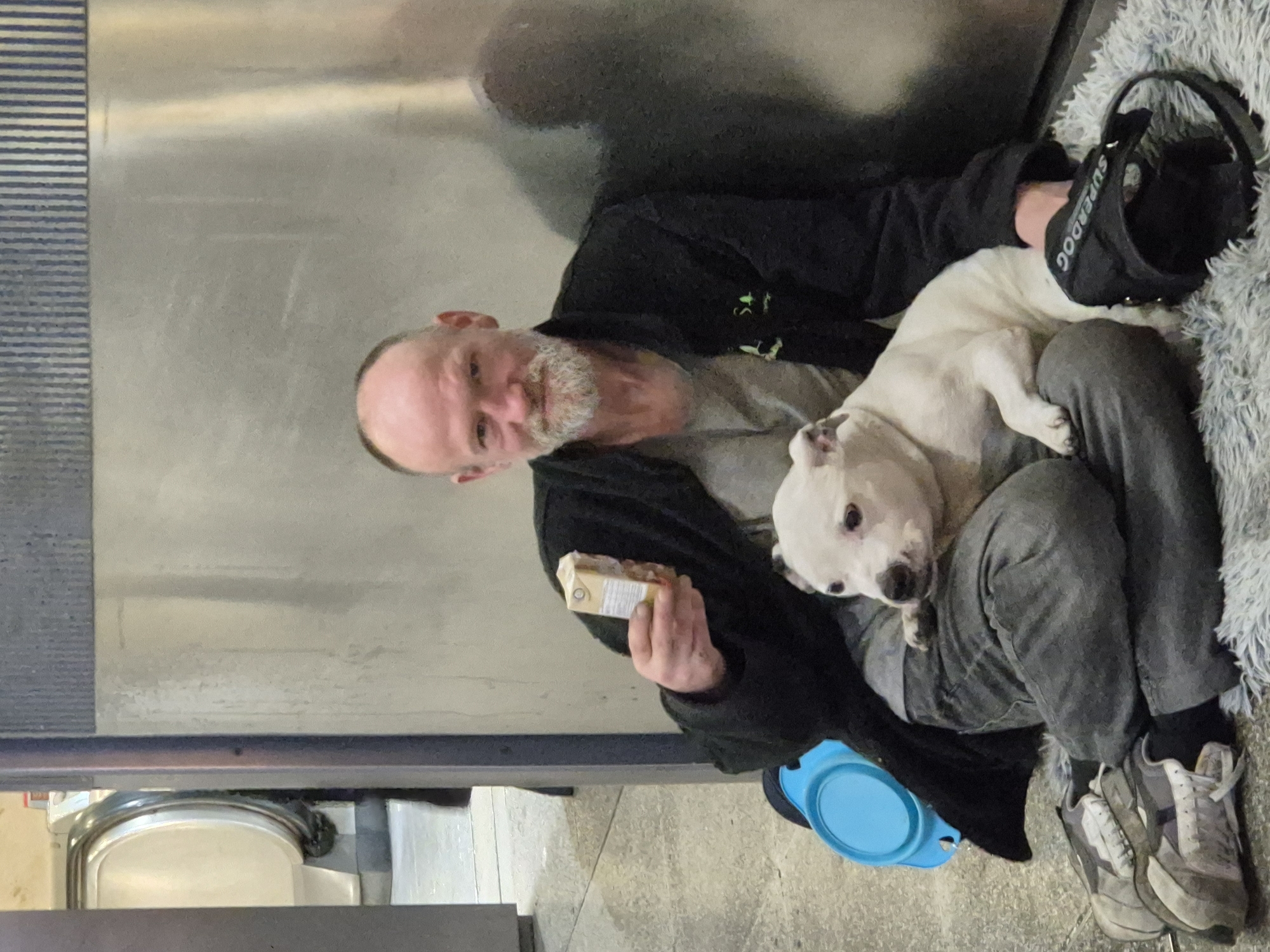 A man with a gray beard sits against a metal wall. He wears a black jacket and gray pants, and gray sneakers. A white dog is curled up on his lap. The man is holding a yellow package. A black hat labeled "Superdog" is beside the dog, along with a gray blanket. A blue bowl is on the floor nearby. The location is likely a building exterior in an urban area. Further detail is indeterminate.