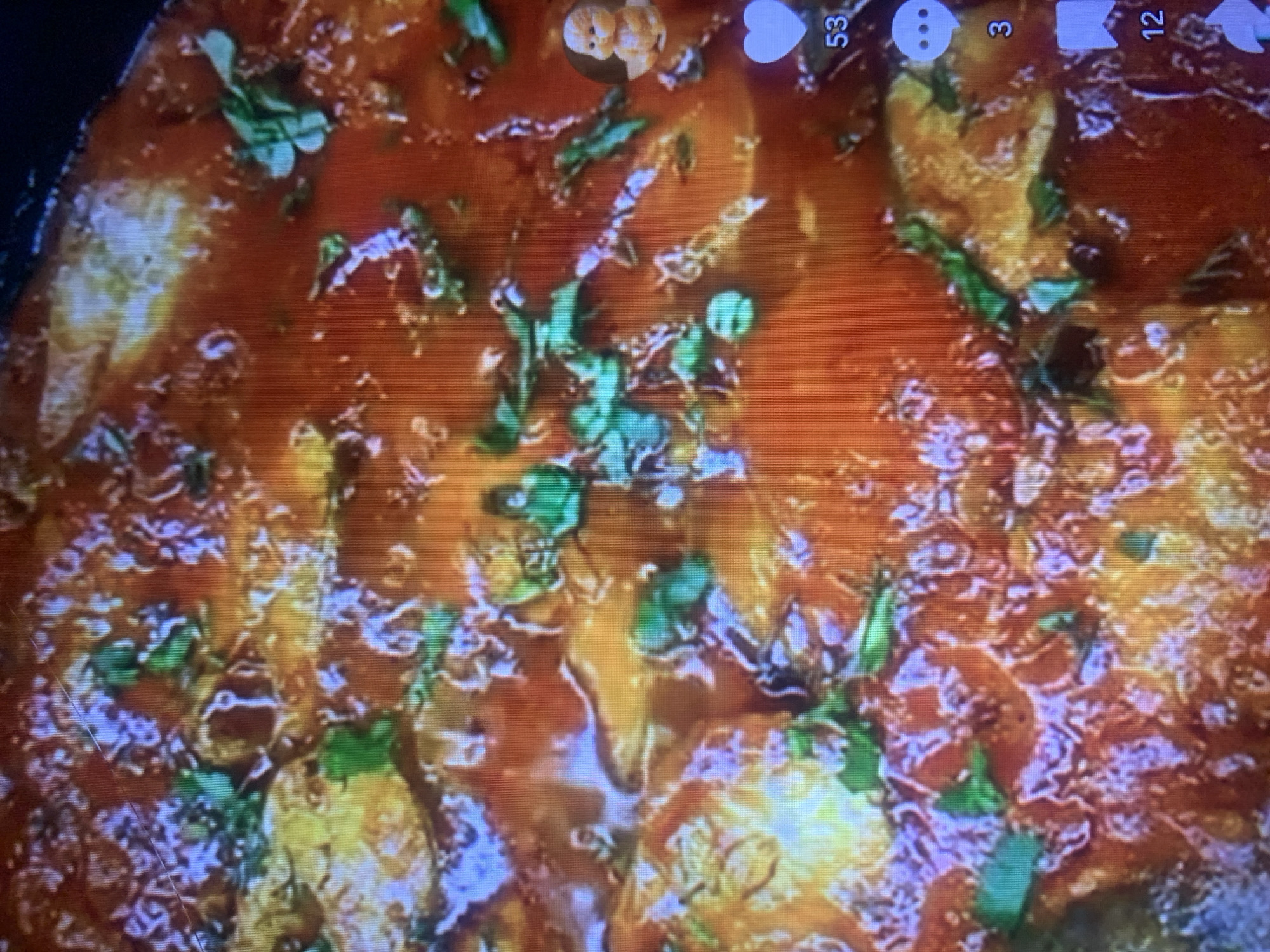 The image is a close-up, overhead shot of a dish of food. The food appears to be pieces of meat in a thick, red sauce, garnished with chopped green herbs. The lighting is warm and creates some glare and reflections on the saucy surface. The image is slightly out of focus and has a grainy texture. There are social media icons on the right side of the image: a baby emoji, a heart icon with the number 53, a speech bubble icon with the number 3, and a bookmark icon with the number 12. The location in the city of Almaty, Kazakhstan, cannot be determined from the image.