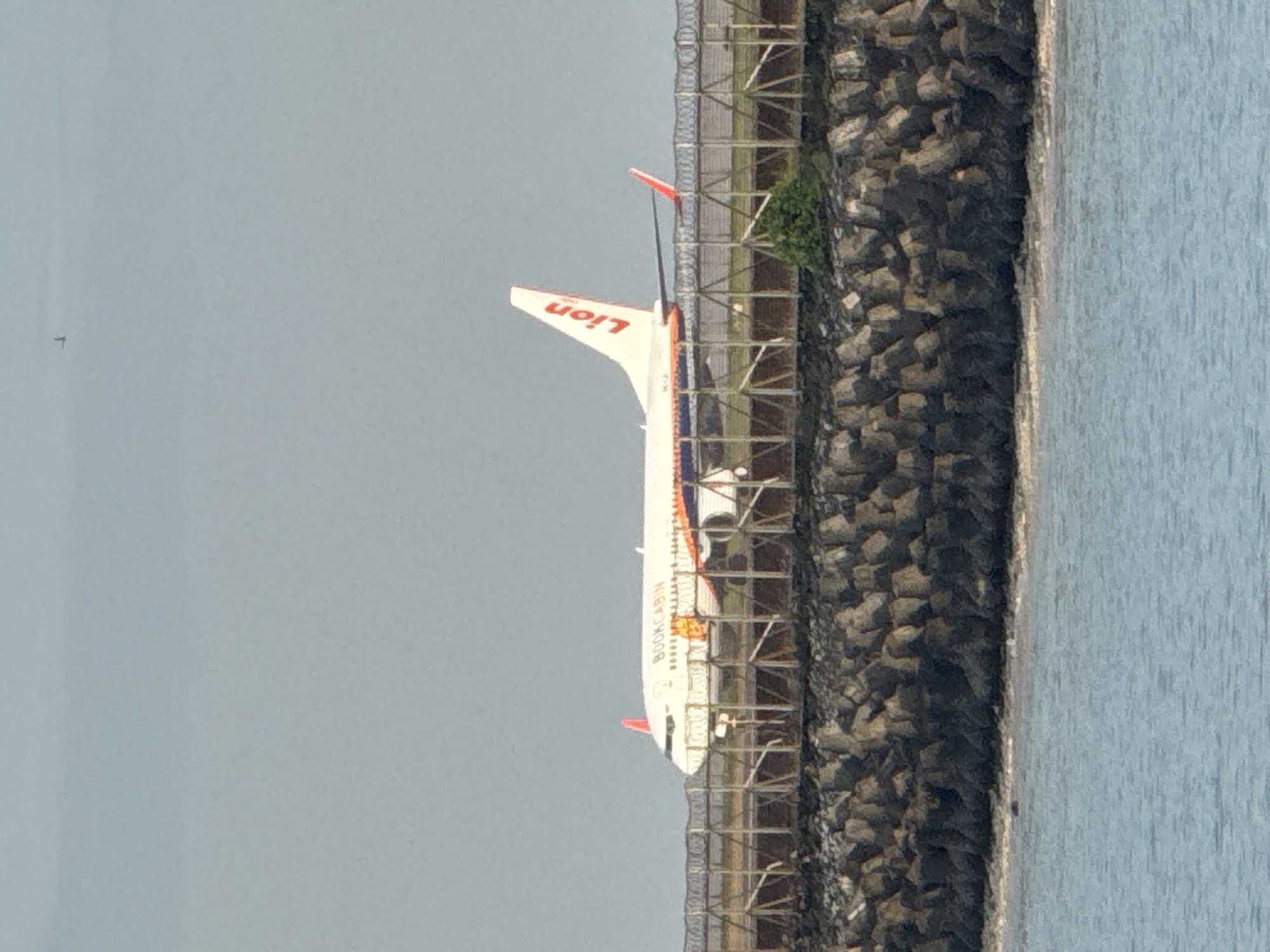 The image depicts a stationary Lion Air Boeing 737-900ER aircraft located on the Ngurah Rai Airport grounds, near the shoreline in Denpasar, Indonesia. The aircraft has the airline's "Lion" logo prominently displayed on its tail. The plane is positioned behind a wire fence and a man-made rocky barrier along the edge of the water. The setting appears to be overcast, with a gray sky and calm water.