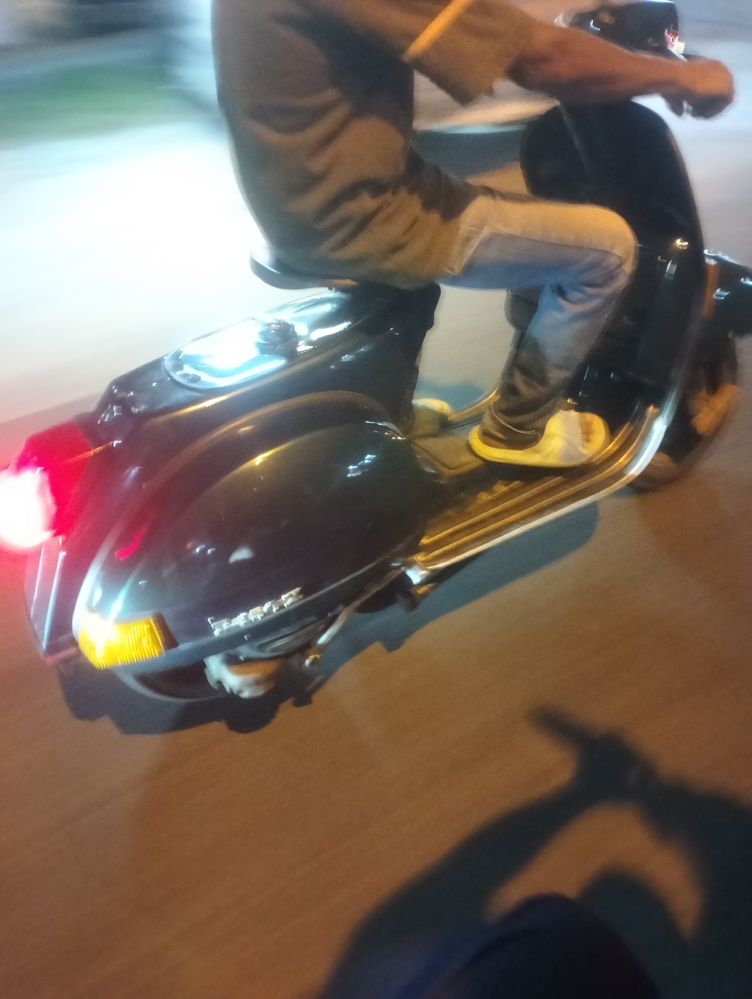 A motion-blurred, nighttime outdoor photograph captured in Duri Barat, Indonesia, shows a person riding a dark-colored Vespa scooter. The rider, only partially visible, is wearing a grey long-sleeved shirt and blue jeans, with yellow sandals on their feet. The scooter's rear brake light is illuminated red, and the front turn signal is a bright yellow. The scooter has a metallic trim along its base. The ground appears to be asphalt, with a shadow of the rider and scooter cast onto it. The background is indistinct due to motion blur, but suggests an urban environment with some illuminated areas.
