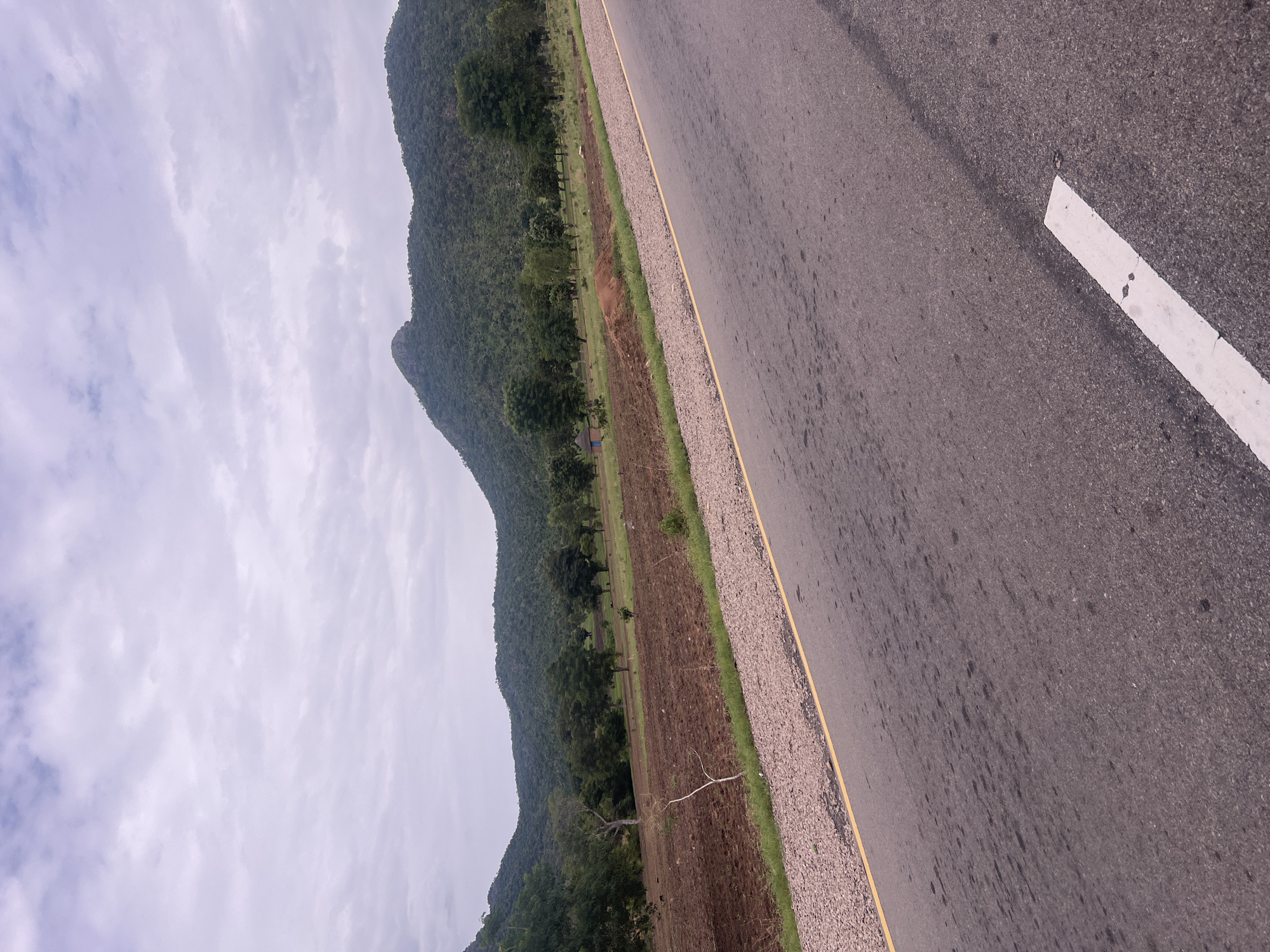 The image shows a rural roadside scene in Minda, Nigeria. The foreground is dominated by a paved road with a white lane marking and a yellow line bordering a gravel shoulder. To the left of the road, a strip of green grass transitions to a field of tilled brown earth, indicating agricultural activity. Further left, a line of trees separates the field from a steep, densely forested mountainside. The sky is overcast with pale, wispy clouds. The scene suggests a cloudy day, likely in the morning or afternoon, given the ambient light. No people are visible, but a small, blue tent is pitched in the field, suggesting a temporary encampment or perhaps a farmer taking shelter. The overall impression is one of a quiet, undeveloped rural landscape.