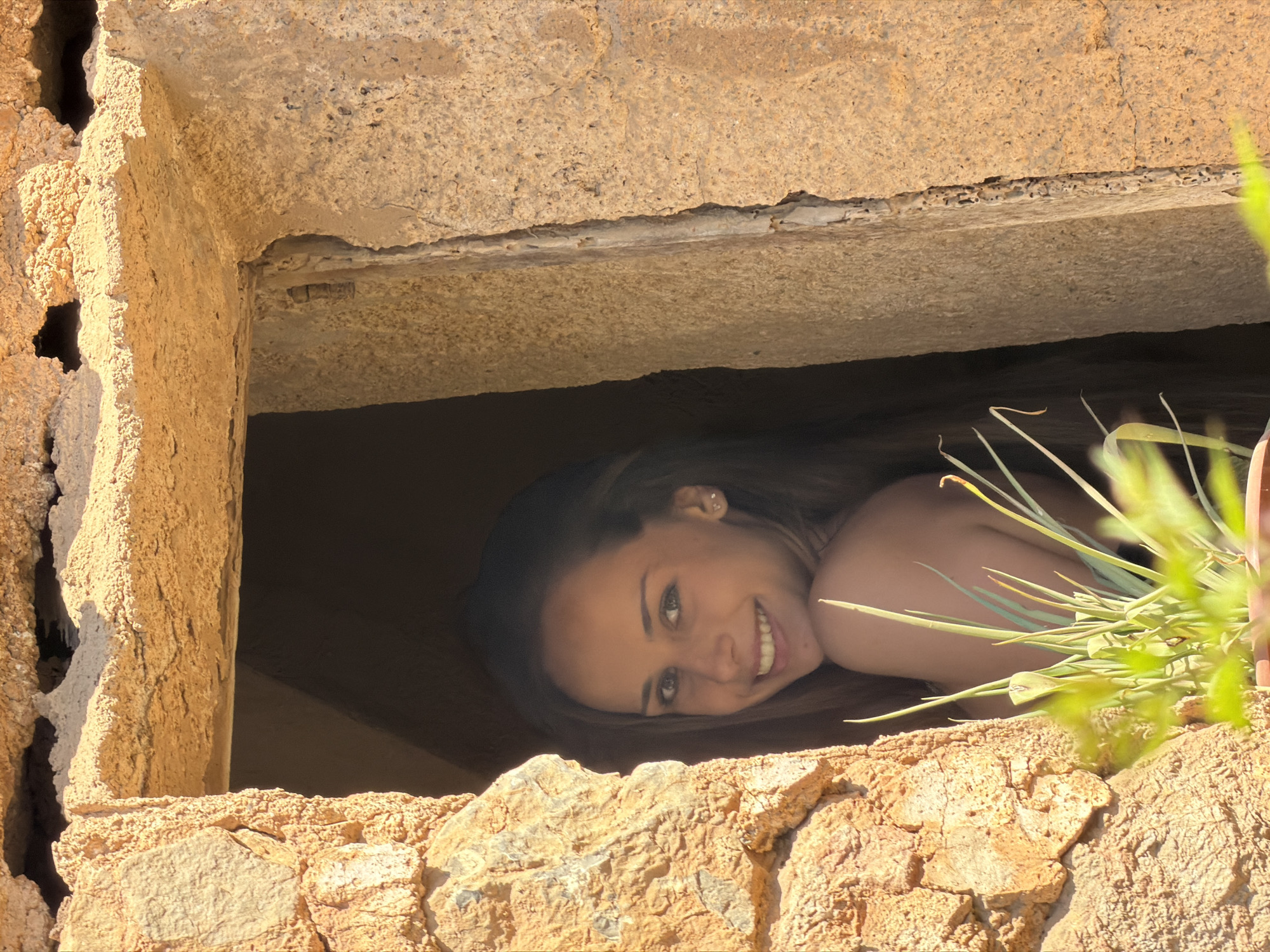 A young woman is looking out from a stone window opening in what appears to be an old stone building. The building is located in Bunyola, Spain, judging by the architectural style. The woman is smiling, and her face is well-lit. Green plants are also seen in the window. The stone structure looks aged and weathered.
