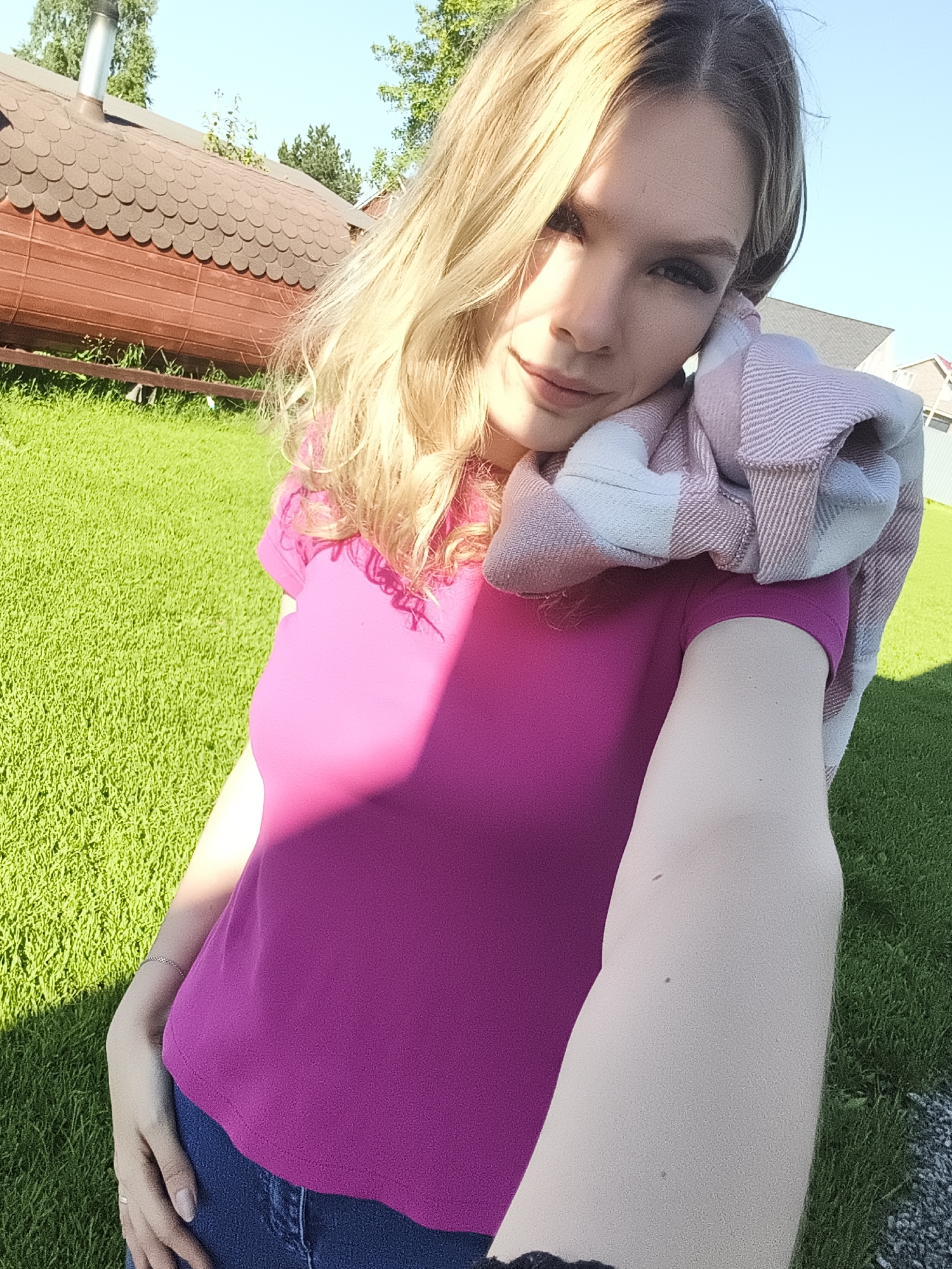 A woman with blonde hair is taking a selfie outdoors in Русский Вожой, Russia. She is wearing a magenta t-shirt and blue jeans, and a pink and white checkered scarf is draped around her neck. The background consists of a vibrant green lawn, a wooden structure with a shingled roof, and trees. The lighting suggests it is a sunny day.