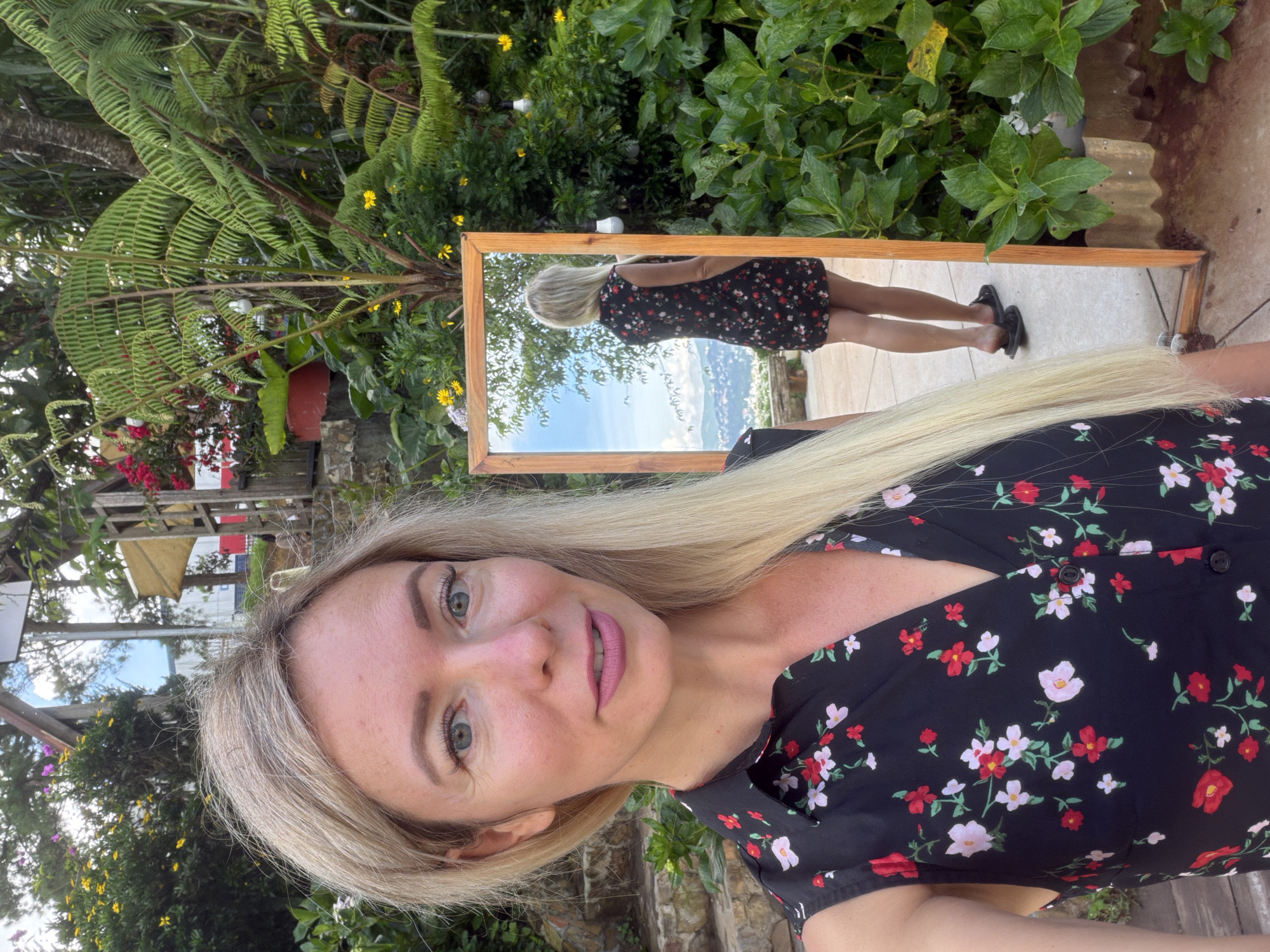 A blonde woman is taking a selfie with a full-length mirror in the outdoor garden of a cafe in Xuân Thành, Vietnam. The woman is wearing a sleeveless black dress with a floral pattern of red, white, and pink flowers. She has light blue eyes and is smiling with pink lipstick on. The mirror reflects her from behind, showing the back of her dress and her legs. She is wearing black sandals. The garden is lush with green foliage, ferns, and a variety of flowers. In the background, there are stone steps, wooden structures, and a view of rolling hills with houses. The sky is visible, with some clouds. The lighting suggests it is daytime, likely late afternoon. The overall scene depicts a relaxed and picturesque setting.