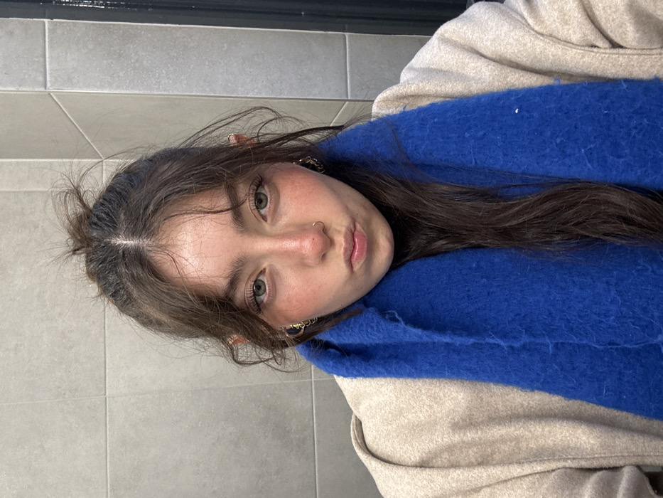 A young woman with fair skin, rosy cheeks, and green eyes gazes at the viewer. Her dark hair is partially pulled back, and she has a nose ring and delicate earrings. She is wearing a beige jacket and a bright blue, fuzzy scarf. The background is a neutral gray, suggesting a tiled wall. The photo is slightly rotated.