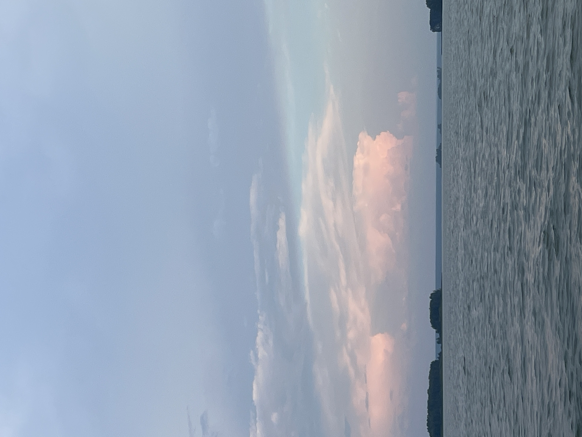 A wide-angle, vertical shot captures a serene lake scene under a pale, muted sky. The water, a mix of grey and blue hues, is gently rippled, creating a soft, textured surface. In the distance, several small, tree-covered islands dot the horizon, appearing as dark green masses against the lighter sky. The sky itself transitions from a pale blue at the top to softer, pastel tones of pink and grey clouds lower down. A large, cumulus cloud formation, tinged with pink, dominates the middle of the sky. The overall impression is one of calm and natural beauty. There are no people or animals visible in the image. The location is the city of Novopichugovo, Russia.