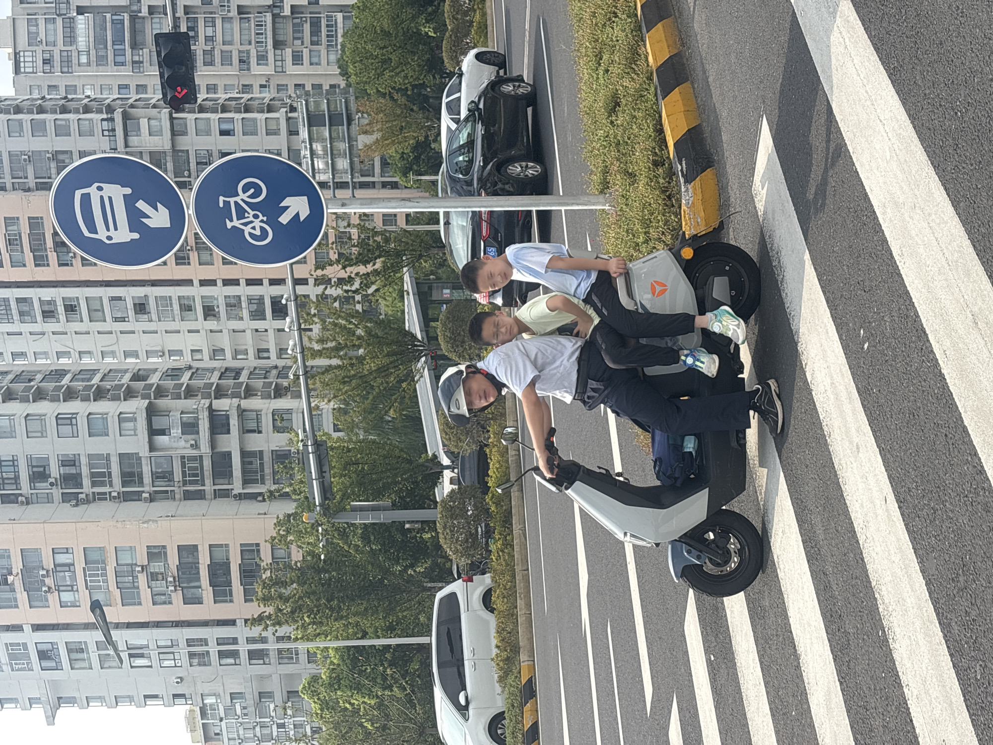 A man wearing a white short-sleeved shirt, black pants, a helmet, and black and white sneakers is riding an electric scooter on a pedestrian crossing. He is seated in the front and appears to be looking forward. Two young boys are seated behind him on the scooter. The boy in the middle is wearing a light yellow short-sleeved shirt, black pants, and blue and white sneakers. The boy behind him is wearing a light blue short-sleeved shirt, black pants, and colorful sneakers. The electric scooter is gray and white with a black seat and a blue bag behind the driver.
The scooter is positioned on a zebra crossing with white stripes. The background shows a wide street with multiple lanes of traffic. Several cars are visible on the road, including a white minivan and a black sedan. To the left of the road, there is a green median strip with bushes and trees. In the background, there are tall, multi-story apartment buildings with many windows, typical of urban development. Traffic lights and street signs are visible above the road. One sign is a blue circular sign with a white car icon and a white arrow pointing down. Below it is another blue circular sign with a white bicycle icon and a white arrow pointing down. A traffic light with red lights illuminated is also visible. The scene is set in the city of Suzhou Industrial Park, China, indicated by the style of the buildings and traffic signs. The lighting suggests it is daytime, likely a clear or partly cloudy day.
