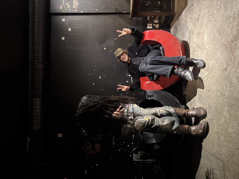 An indoor scene in an unidentifiable venue within Paris, France, features two individuals. The environment is dimly lit, with a dark, distressed wall displaying faint markings and peeling paint as the backdrop. An exposed dark ventilation pipe runs horizontally across the upper portion of the wall. The floor is a light-colored concrete or similar hard surface.
On the left, an individual with long, dark dreadlocks is standing, mostly facing away from the camera or with their head lowered, obscuring their face. They wear a dark jacket, light-colored jeans with multi-colored paint splatters or patterns, and dark brown, worn leather boots with buckles. Their hands are not clearly visible.
On the right, another individual is seated in a striking red, U-shaped armchair, directly facing the camera. This person wears a camouflage-patterned baseball cap, a dark velvet or corduroy blazer over a dark top with a silver chain and pendant, dark trousers, and black pointed boots featuring multiple buckles and studded details. They are smiling broadly and holding up both hands, forming a "peace" (V-sign) gesture with each. Their legs are crossed at the ankles.
Adjacent to the red armchair, to its left, is a similar U-shaped structure, black in color, which is unoccupied. A light-colored wooden table is partially visible on the far right of the frame.