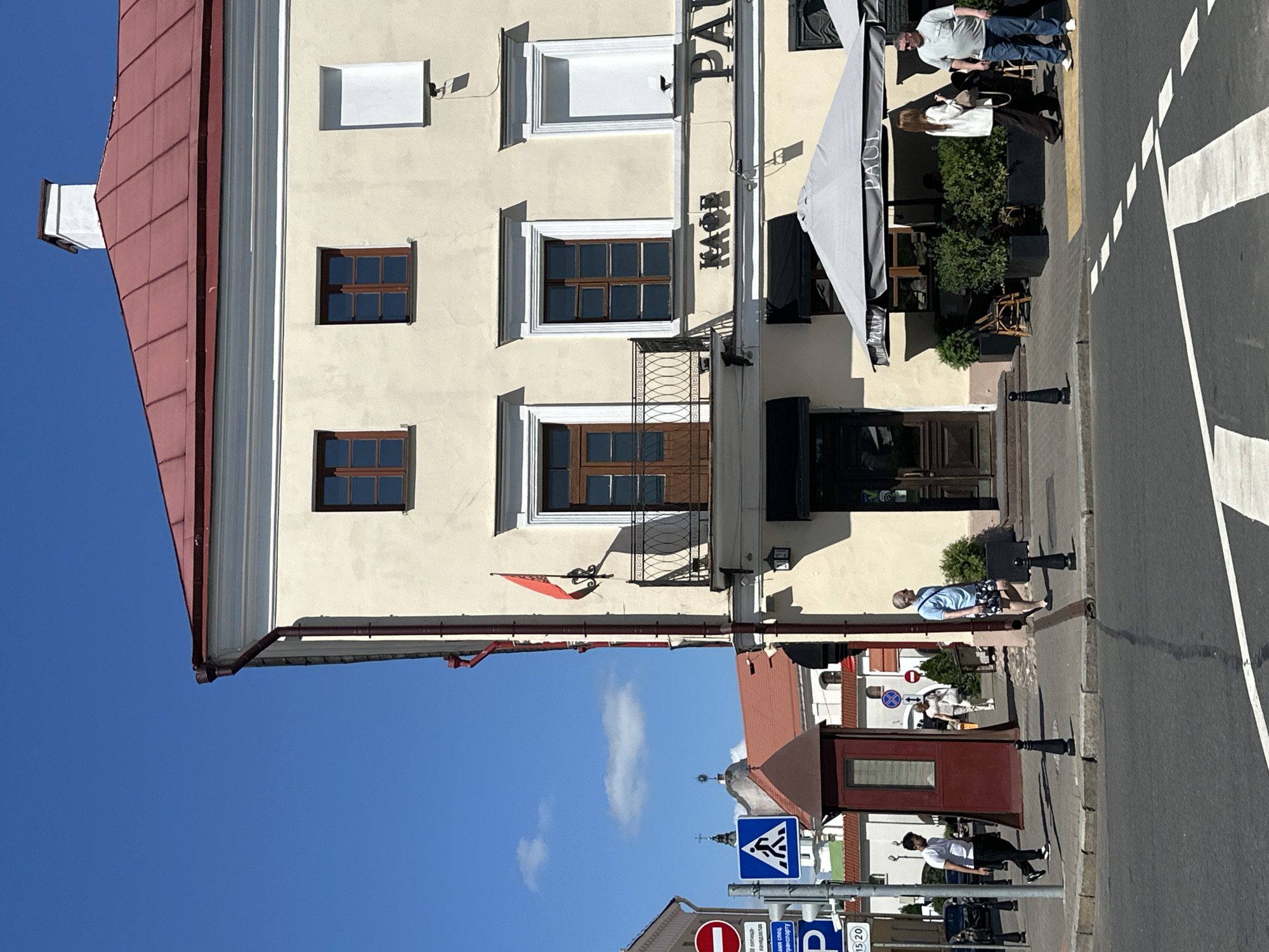 The image shows a street scene in Minsk, Belarus, on a sunny day with a clear blue sky. A multi-story building with a red roof and cream-colored walls dominates the right side of the frame. There are several windows on the facade, some with wooden frames and others with white shutters. A balcony with an ornate railing is visible on the second floor, with a wooden door leading onto it. A small red flag is displayed on a decorative bracket on the wall.
Below the balcony, the building features signage that reads "KAФE" and "PAUL". An outdoor seating area for the establishment is set up under a large grey umbrella with "PAUL" written on it. Several people are visible on the sidewalk.
On the left side of the street, there are various street signs, including a red circular sign with a white bar and a blue square sign depicting a pedestrian crossing. A maroon-colored kiosk or booth stands on the sidewalk.
Several individuals are walking on the sidewalk. One man in a white t-shirt and black pants is walking towards the left. Further down, a woman in a light-colored outfit is walking towards the right, and a man in a blue t-shirt and patterned shorts is walking towards the right. Another couple, a woman in a white blazer and dark pants, and a man in a grey t-shirt and blue jeans, are standing near the outdoor seating area.
The street in the foreground has a pedestrian crossing marked with white lines. Cars are parked or driving on the street in the background. The architecture suggests an urban environment.