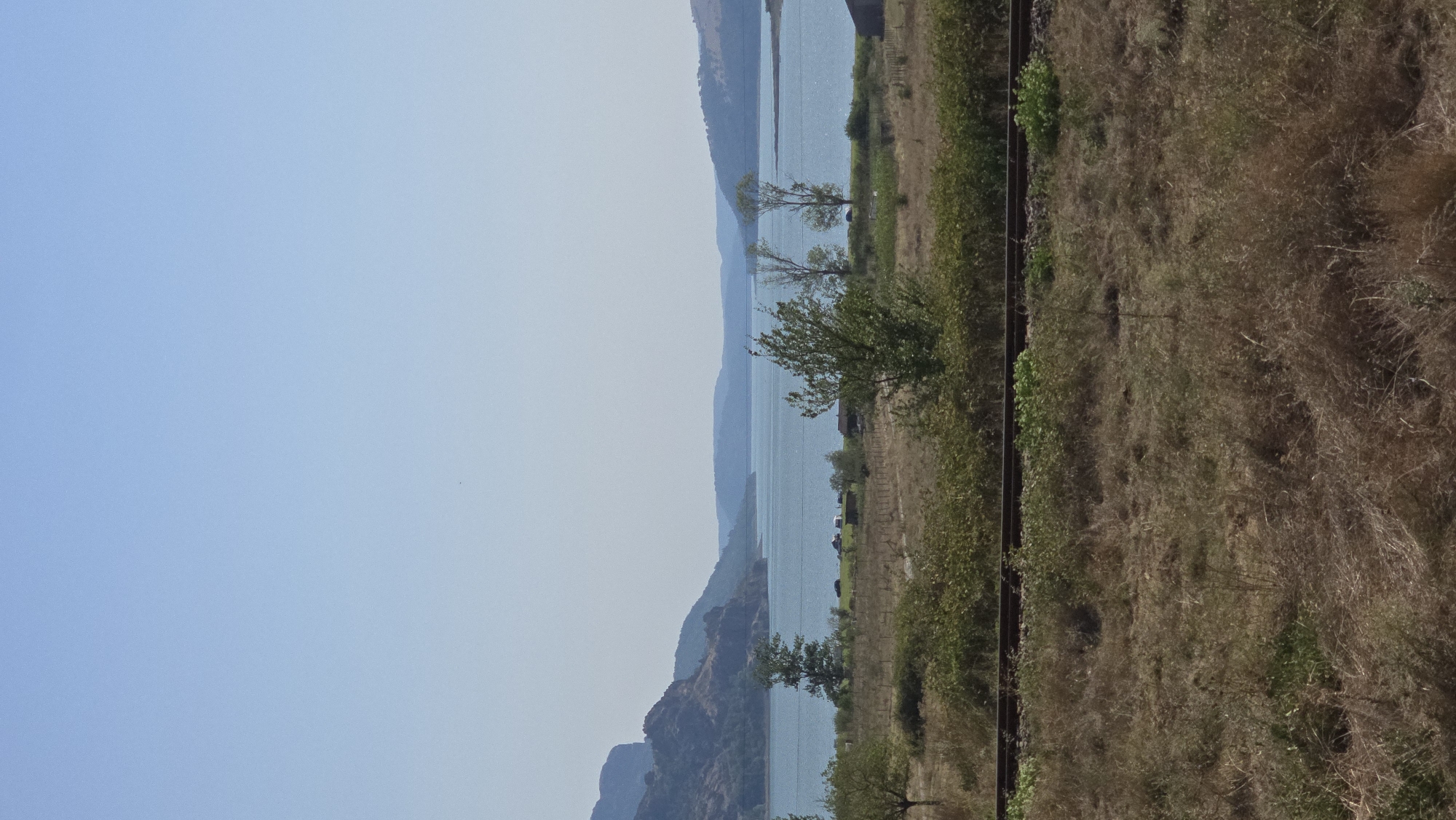 The image depicts a wide-angle, landscape view of a serene body of water, likely a reservoir, with rolling hills and mountains in the distance. The sky is a clear, pale blue, suggesting a sunny day. In the foreground, there is a dry, grassy field with scattered bushes and a section of railway tracks running horizontally across the frame. Some trees are visible, with one prominent tree with sparse leaves positioned near the center of the mid-ground. In the distance, along the shoreline, there are a few small structures and vehicles, hinting at a settlement or recreational area. The overall scene is peaceful and natural, with a sense of open space.