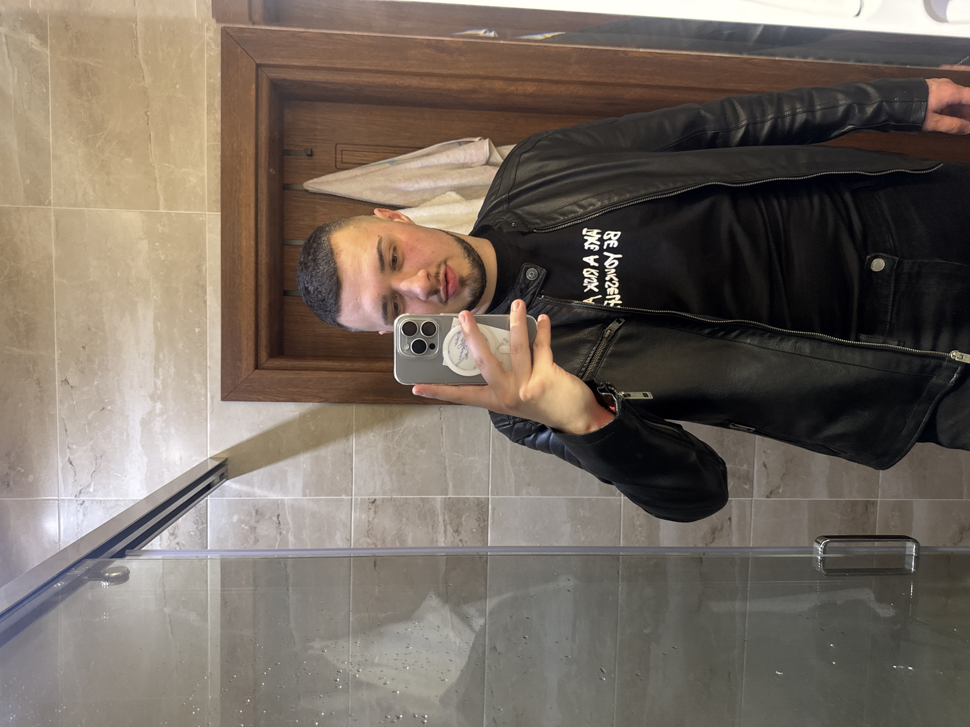 A man is taking a selfie in a bathroom. He is wearing a black leather jacket and a black T-shirt with white lettering. He is holding a silver iPhone. A towel is hanging on a rack in the background. The bathroom has beige marble tiles and a glass shower door. The location is not possible to be determined.