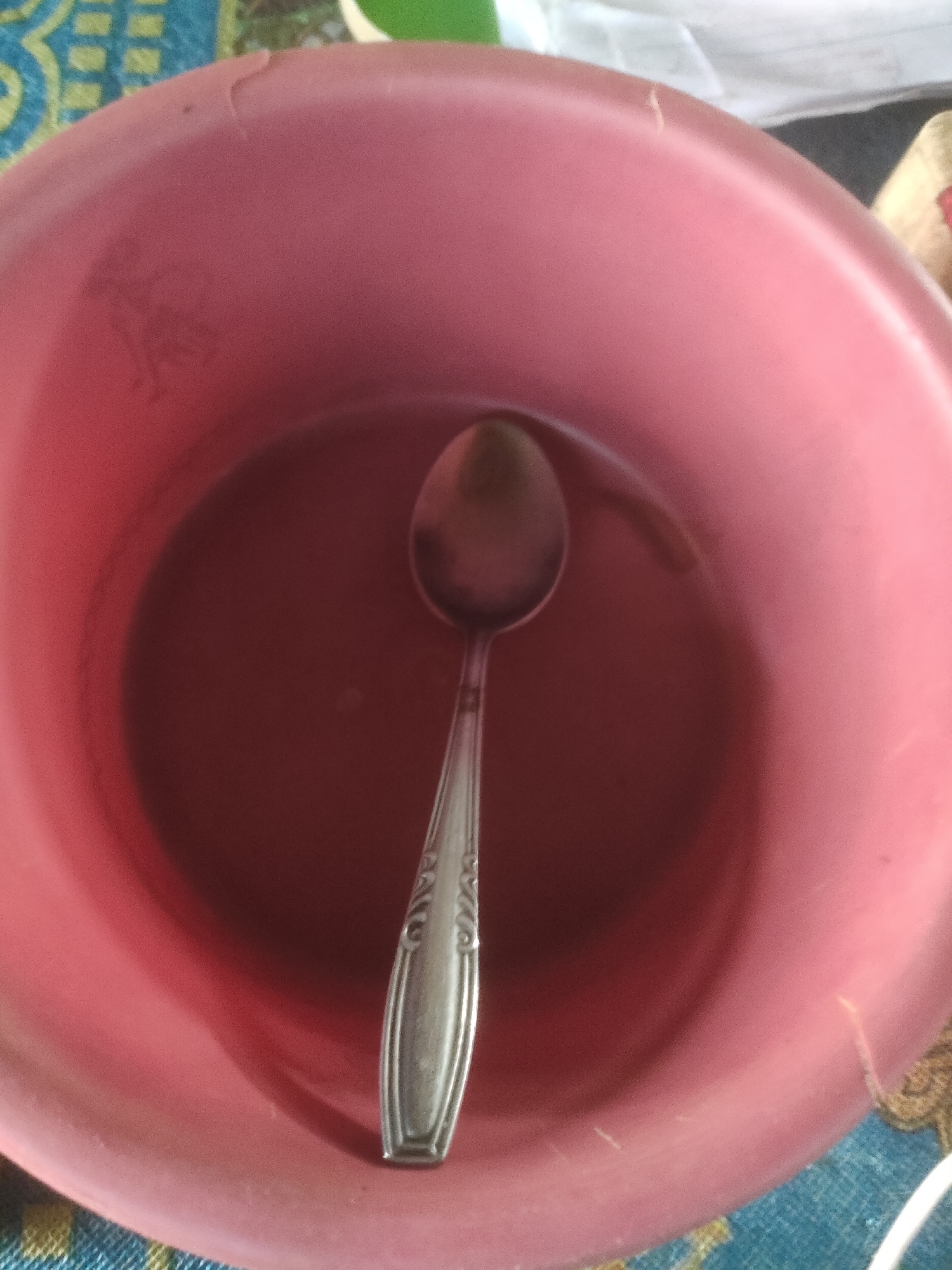 In the city of Padang Dalom, Indonesia, a close-up, overhead shot captures a pink plastic bowl containing a reddish-brown liquid, likely tea or coffee. A silver-colored spoon rests submerged in the liquid, its handle extending upwards towards the rim of the bowl. The bowl is situated on a surface covered with a patterned cloth, featuring shades of blue and yellow. The lighting is dim, creating shadows within the bowl and highlighting the texture of the plastic. The image is a still life, focusing on the simple arrangement of household items.
