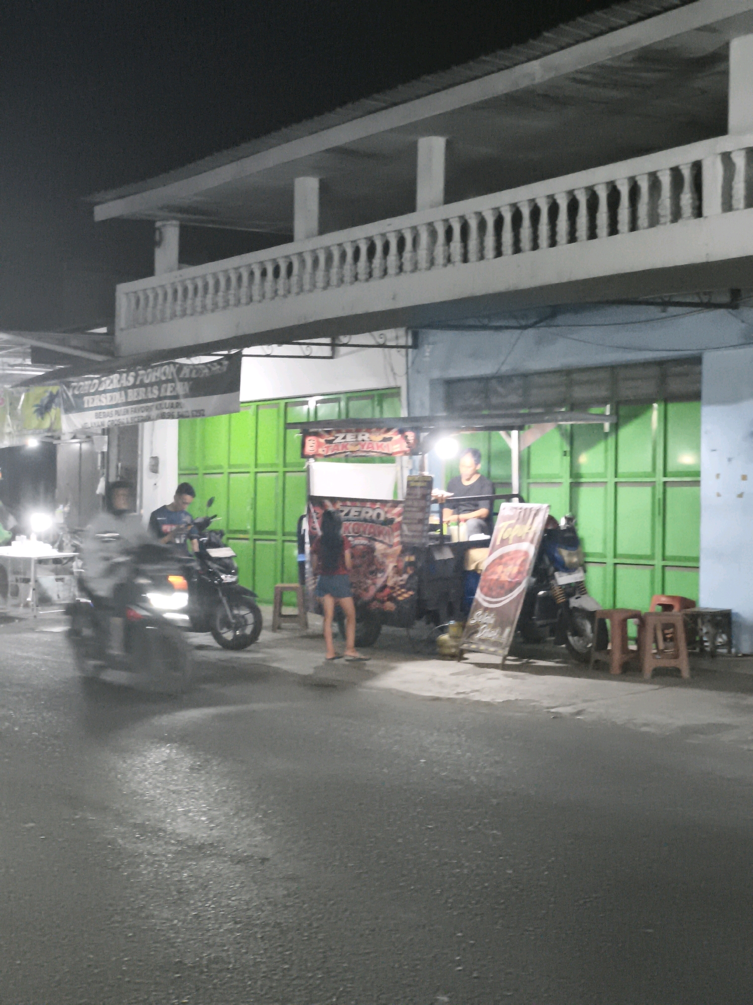 This image captures a nighttime scene on a street in Desa Donohudan, Indonesia, likely a commercial area. In the foreground, a street is wet, reflecting the ambient light from nearby establishments. The middle ground shows a food stall with green metal shutters behind it. A person wearing a dark t-shirt is standing next to a motorcycle in front of the stall. A woman in a blue skirt and dark top is also visible near the stall, and two small, light-colored stools are placed on the ground. A large banner, partially obscured and blurry, is displayed on the stall, featuring text and images, including the word "ZERO." Another motorcycle, with a rider, is in motion on the left side of the frame, appearing blurred due to movement. Further left, a white table with chairs is set up, illuminated by a light source. Above the stall and street, an elevated structure with a white railing and pillars is visible, suggesting it's an overpass or a second story of a building. A dark banner with white text, possibly advertising goods, hangs from the elevated structure. The overall atmosphere is dimly lit, characteristic of a street scene at night.