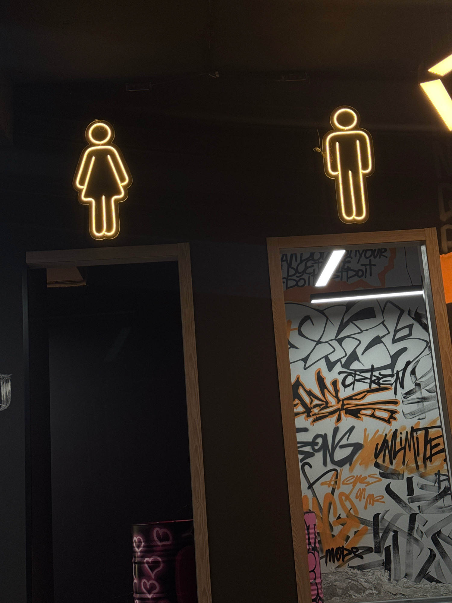 The image shows two neon gender signs, one depicting a female figure and the other a male figure, illuminated against a dark background. The female sign is positioned above the entrance to a dark doorway on the left, while the male sign is above a doorway on the right. The doorway on the right is open, revealing a wall covered in graffiti, predominantly in black, white, and orange. The graffiti includes various tags and artistic designs. A bright fluorescent light fixture is visible inside this doorway. A partially visible metal barrel with neon pink heart shapes is also present inside the dark doorway on the left. The overall setting appears to be an indoor establishment, possibly a restroom area within a commercial or entertainment venue. There are no people visible in the image. The location is not specified within the image itself.
