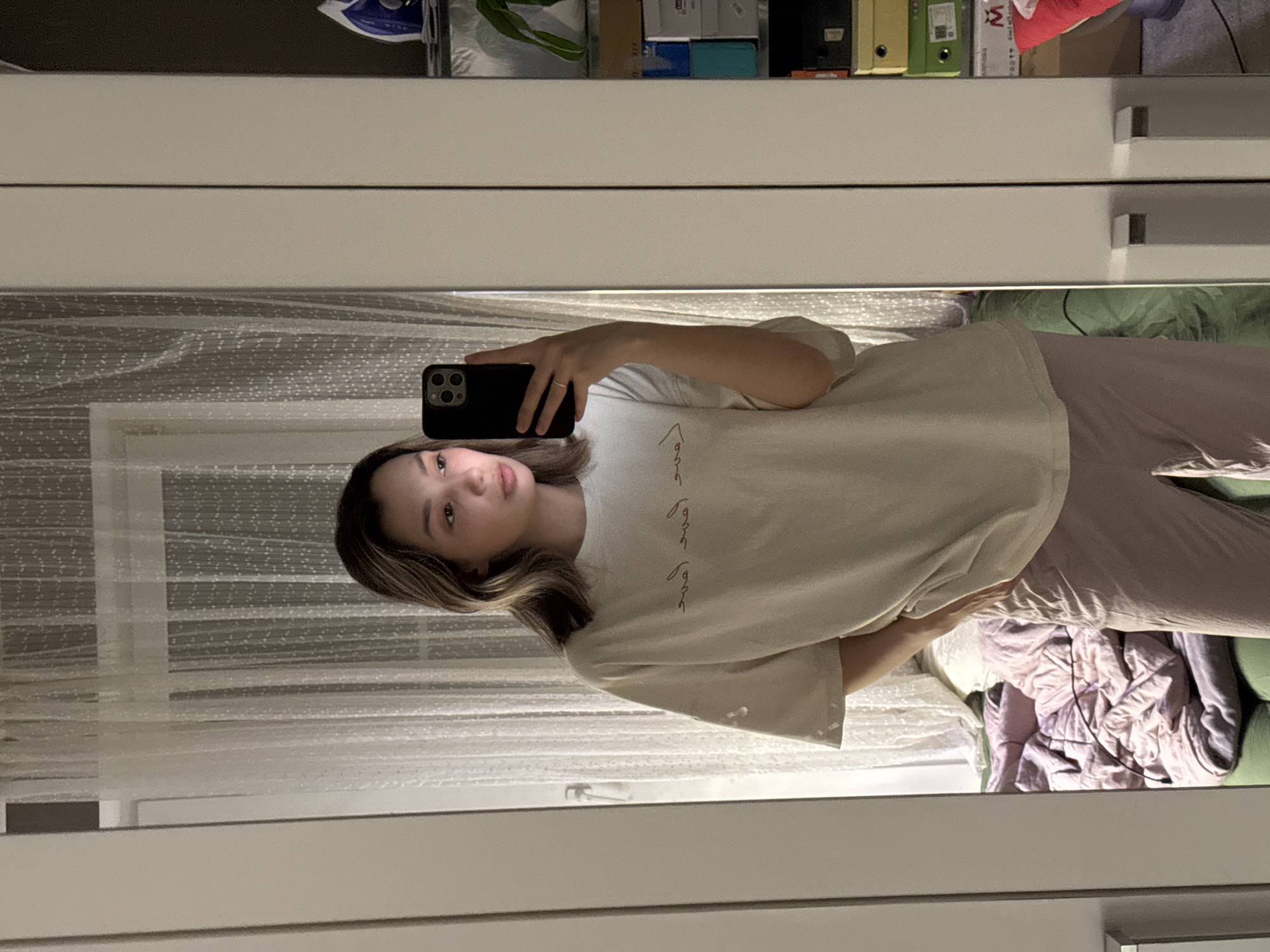 The image shows a young woman or girl taking a mirror selfie indoors. She holds a black smartphone, likely an iPhone with a triple camera setup, in her left hand, which partially obscures her face. She has medium-length, highlighted hair and a neutral expression, looking towards the phone's reflection. She is wearing an oversized, light-colored (possibly off-white or cream) t-shirt that features faint, stylized red or brown script on the front. Her lower body is clad in light-colored, loose-fitting pants. A simple ring is visible on her left ring finger.
The setting appears to be a bedroom. The mirror reflects a bed with crumpled bedding in shades of green and purple or pink. Behind the person, a white, translucent curtain with a subtle textured pattern suggests a window. Above the mirror's reflection, there is a white cabinet or shelf unit holding various items, including boxes, folders, and what appears to be an iron. The lighting is soft. No specific time of day or weather is discernible.