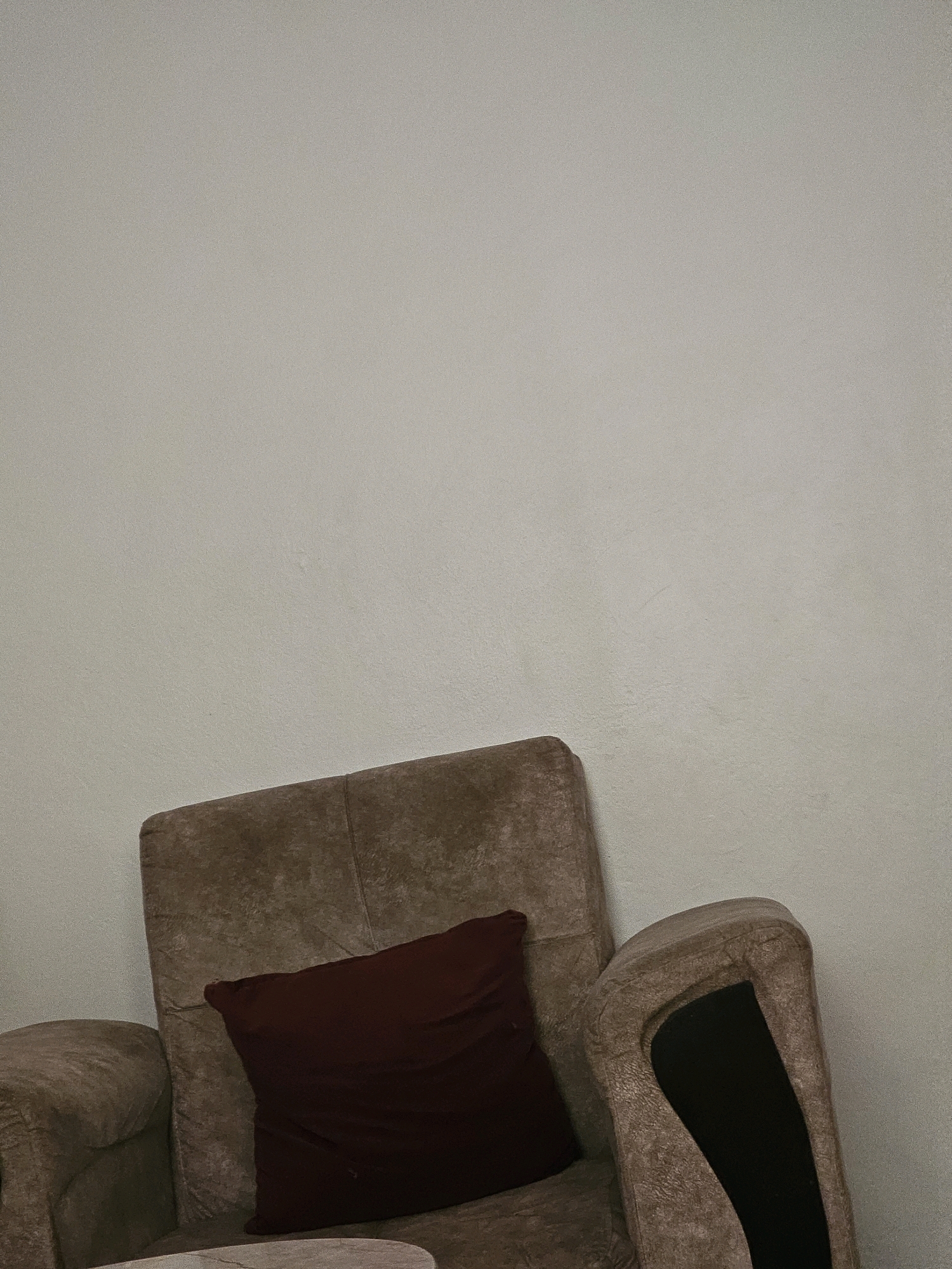The image shows a close-up of a brown armchair with a dark red cushion against a plain, light-colored wall. The armchair appears to be upholstered in a textured fabric, possibly velvet or suede, and has a rounded armrest. The wall is smooth and free of any noticeable decorations or features, suggesting a simple interior setting. The lighting in the image is soft, casting subtle shadows that highlight the texture of the armchair and cushion. No people are visible in the image. The scene is static and peaceful. The image does not provide any information about its location in Batna, Algeria or any specific context.