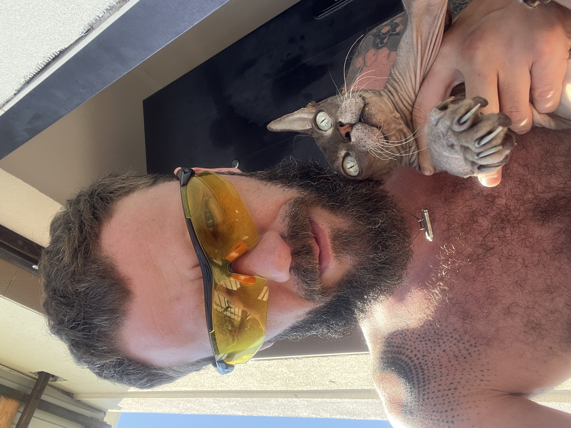 A man with a beard and yellow-tinted sunglasses is holding a Sphynx cat in front of a light-colored wall and a dark structure. The man is shirtless, revealing tattoos on his chest and arm. The cat has green eyes and is looking upwards. The scene appears to be outdoors, possibly in an urban or suburban setting, given the architectural elements. The overall impression is a candid snapshot, possibly taken during daytime. The location in Балаклавский округ, Ukraine is not determinable from the visual information.