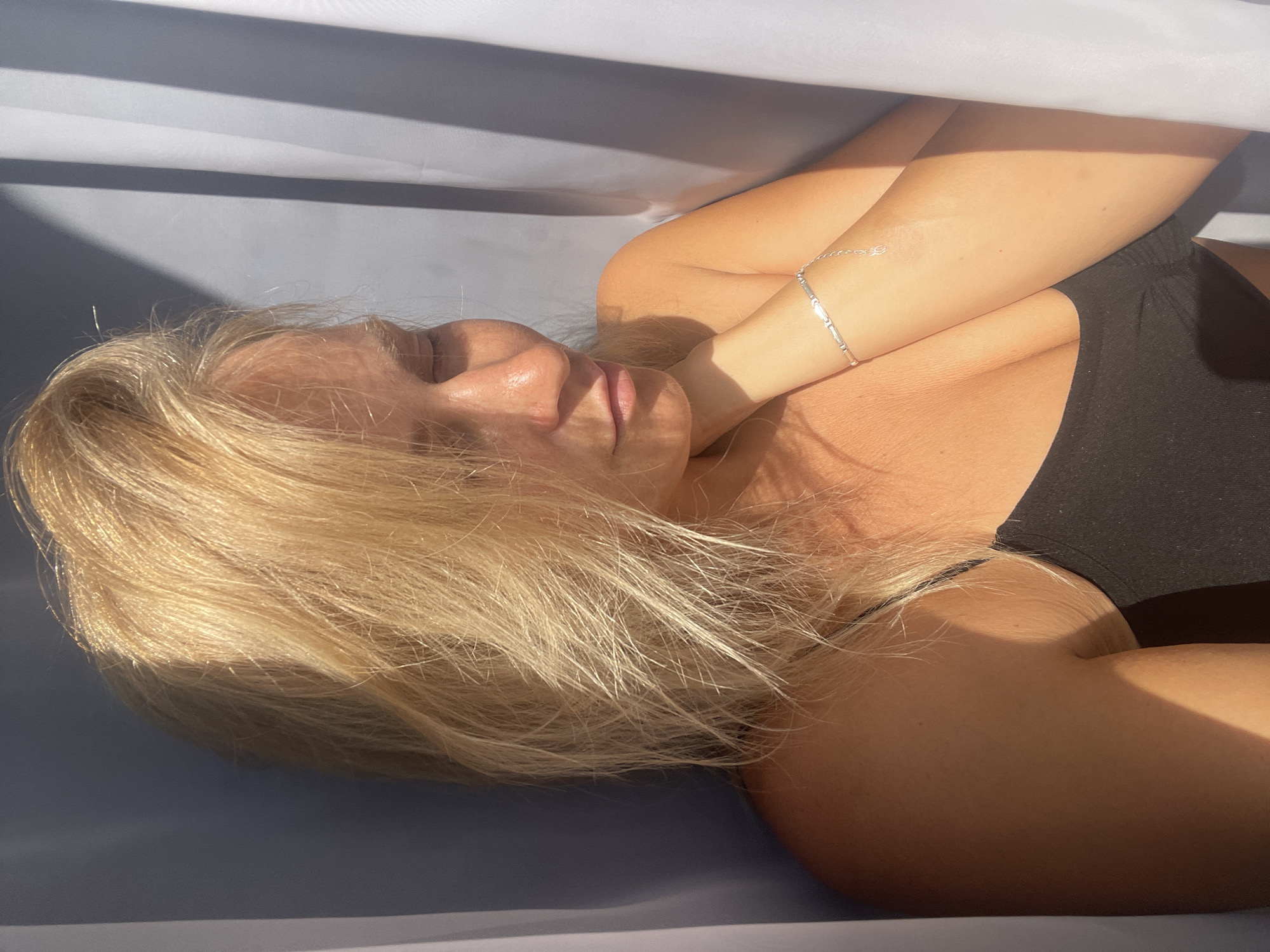 A medium shot captures a fair-skinned woman with blonde hair, her eyes closed, tilting her head to her right shoulder as she touches her neck with her left hand. She is wearing a black tank top and a silver bracelet on her left wrist. The lighting suggests she is near a window, with sunlight casting shadows across her face and upper body, creating a warm glow. The background is a draped fabric, with a darker shade on the left and a lighter, sheerer shade on the right, partially obscuring the wall behind. There is no information available to determine the location in the city of Baranovka, Russia.