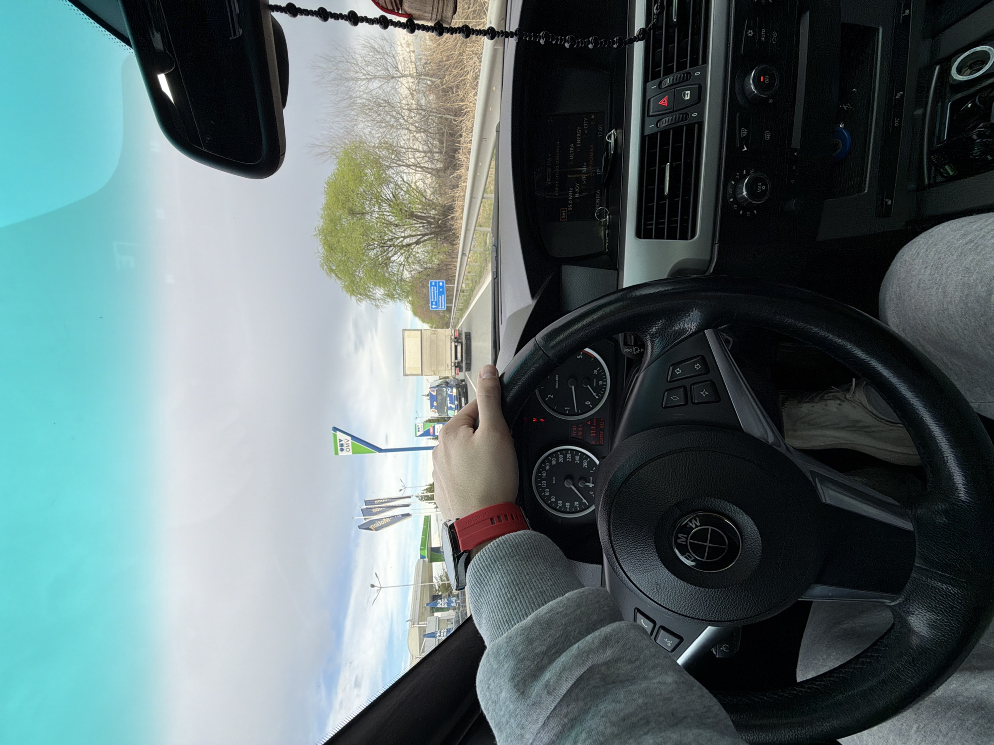 The image depicts the interior of a BMW car, specifically the driver's side, with a person holding the steering wheel. The person wears a grey hoodie and a red smart watch. The car's dashboard and controls are visible, including the instrument cluster with speedometer and other gauges, the radio display showing "95.0 MHz" and station names like "N-JOY", "ENERGY," and "CITY," and the climate control system. Outside the car, through the windshield, is a road with other vehicles, a green tree, and signs indicating a gas station. The gas station brand is "OMV" and it's located in Plovdiv, Bulgaria, suggested by the Cyrillic text "noitom" which is the mirror-image of "Mototoin" a fuel distributor located in Plovdiv, Bulgaria.