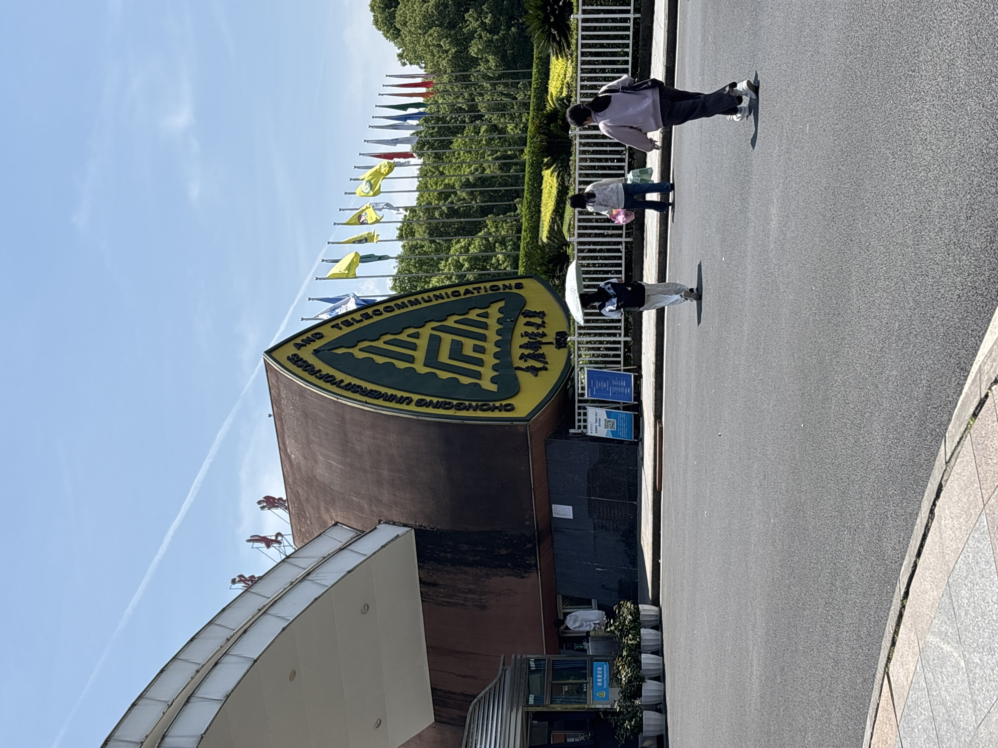 A sunny outdoor scene captures the entrance area of what appears to be a university or institutional campus. On the right side of the frame, a large, curved, reddish-brown building stands prominently, adorned with a distinctive green and yellow triangular sign. The English text on this sign clearly identifies the institution as "CHONGQING UNIVERSITY OF POSTS AND TELECOMMUNICATIONS," accompanied by Chinese characters.
Behind the building and extending across the mid-ground are numerous flagpoles flying various colored flags, backed by dense green trees and foliage. The sky above is a clear blue, featuring a distinct white contrail from an aircraft.
In the foreground, an asphalt road or pathway runs from left to right, where three individuals are seen walking. The person closest to the camera is wearing a light purple top and dark pants, carrying a dark shoulder bag. Ahead of them, another individual in a light top and jeans carries a pink bag. The furthest figure wears a dark top and light pants, holding a white umbrella. To the left of the road, a white fence or railing separates the pathway from green shrubs. Near the building's entrance, two blue information signs are visible; one contains a QR code, and another likely indicates "Visitor Registration" in English and Chinese.
The bright sunlight and clear shadows suggest it is either morning or afternoon. The overall impression is one of a busy, well-maintained campus entrance in Nan'an District, China.