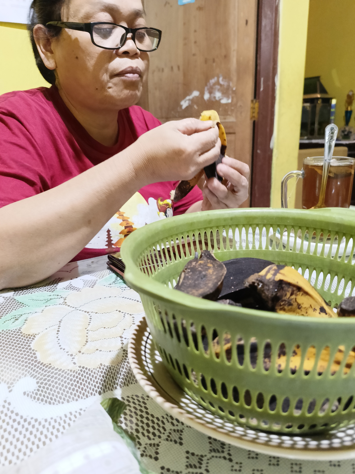 A medium shot shows a person wearing glasses and a red t-shirt, peeling a banana over a green plastic basket filled with banana peels. The person is seated at a table covered with a white lace tablecloth with a floral pattern. A mug of tea with a spoon is visible in the background, along with a wooden door and yellow wall. The scene is indoors, in what appears to be a home in Sukorini, Indonesia. The lighting is warm and artificial.
