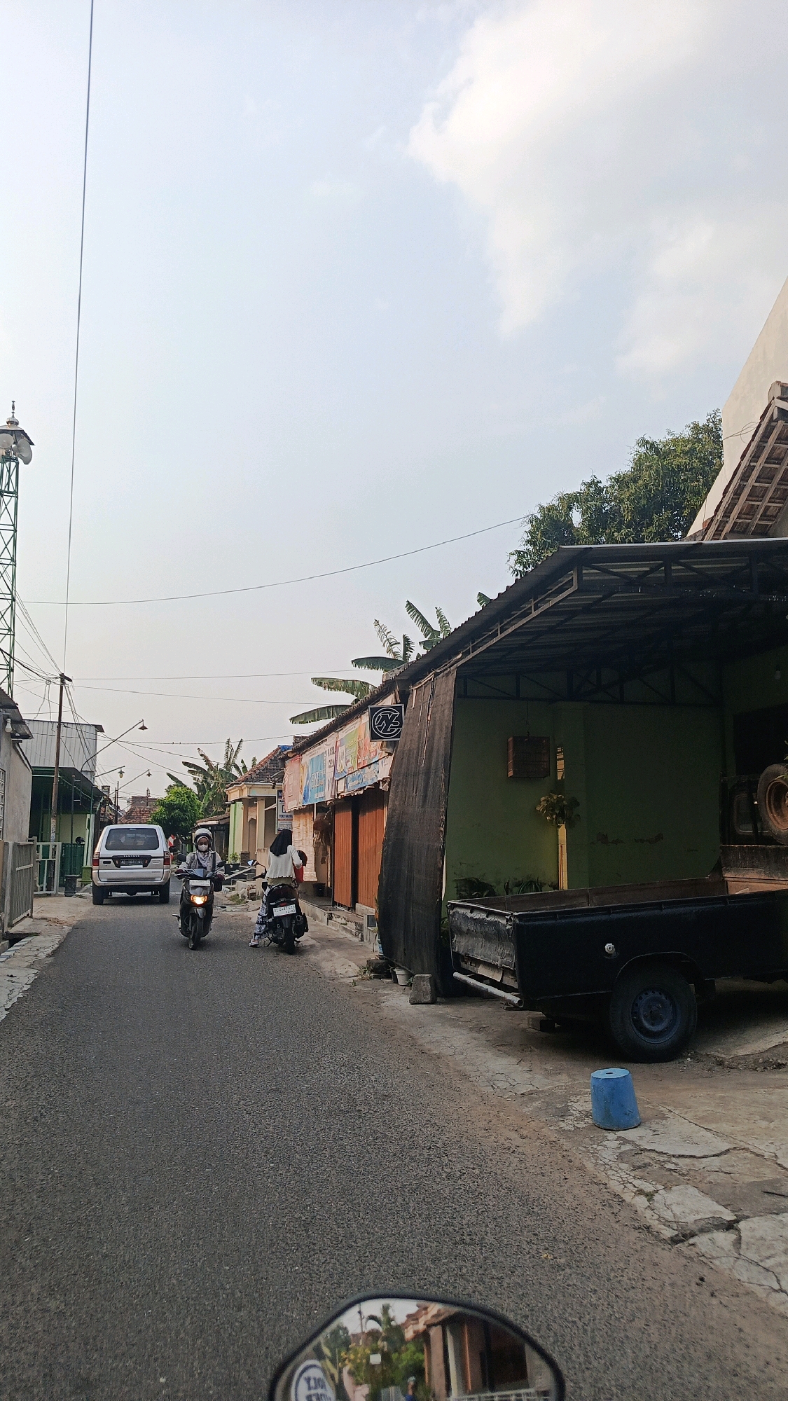 The image depicts a street scene in Mangunsuman, Indonesia, captured from the perspective of someone riding a motorcycle, as indicated by the motorcycle's rearview mirror in the foreground. The street is paved and appears to be a two-lane road.
On the left side of the street, there is a silver SUV parked. Two motorcycles are in motion on the road. The first, closer to the foreground, is ridden by a person wearing a white headscarf and a light-colored top. The second motorcycle, further down the road, is ridden by a person wearing a white top and darker pants.
The right side of the street is lined with buildings, appearing to be shops or residences. These buildings have varying architectural styles, with some featuring corrugated metal roofs and wooden storefronts. One building is painted a distinct green and has a large, dark tarp covering part of its facade. A black pickup truck is parked beside this green building. Further down the street, more buildings with orange doors and various signs are visible.
Overhead, power lines crisscross the sky, and a tall communication tower with multiple antennas is visible on the far left. The sky is overcast with a light haze. Lush green trees and foliage are interspersed throughout the scene, suggesting a semi-urban or rural environment. The overall impression is a typical street in a town or village in Indonesia during daylight hours.
