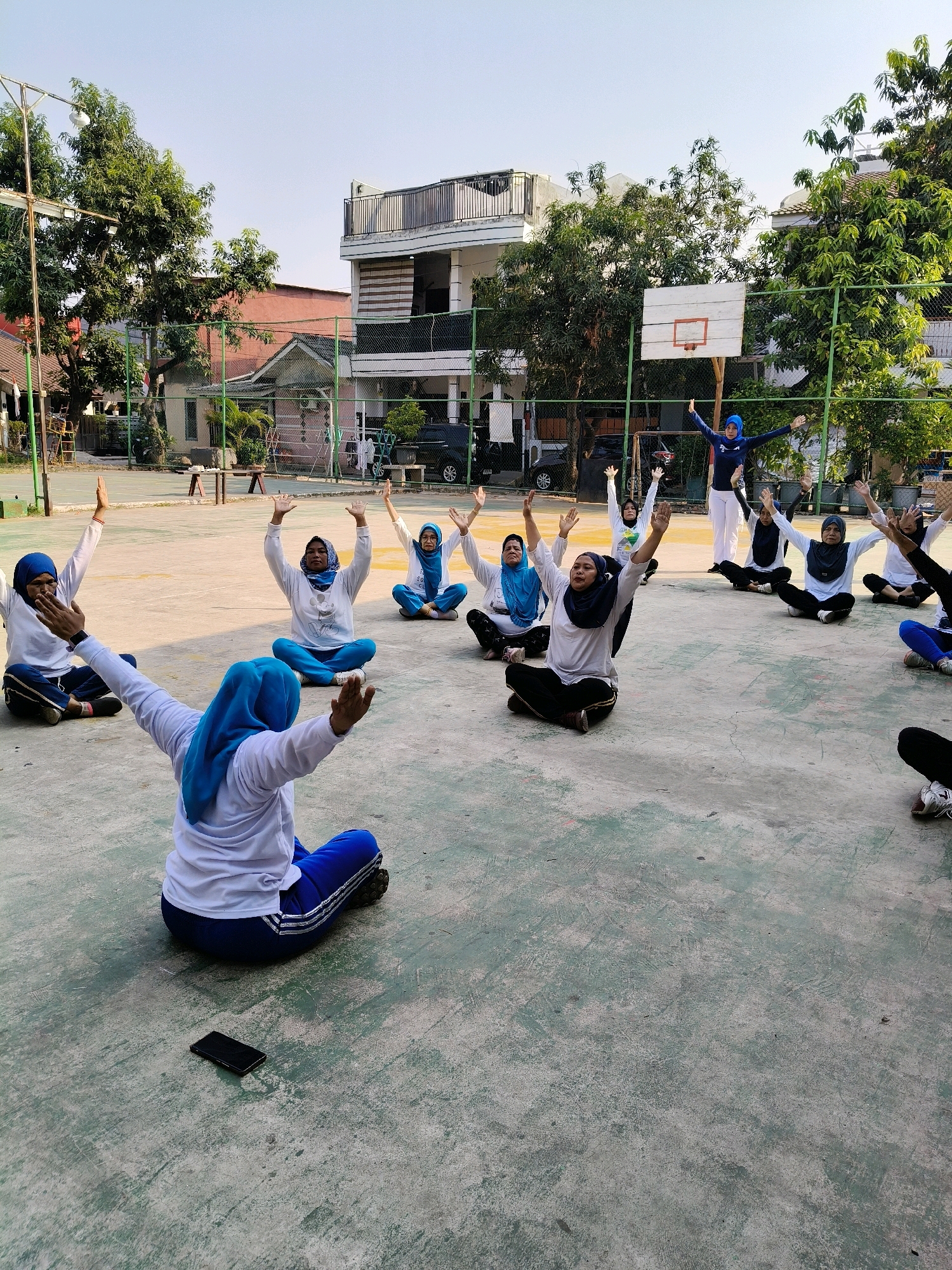 In Setiamekar, Indonesia, a group of women, most wearing hijabs and athletic attire, are participating in an outdoor exercise class on a paved court. They are arranged in a semi-circle facing a woman standing and gesturing. The standing woman, also in athletic wear and a hijab, appears to be leading the session. The seated women are positioned with legs crossed, some with arms raised, suggesting they are following instructions. The court is surrounded by a chain-link fence, with trees and buildings visible in the background, including a multi-story building and a basketball hoop. The ground surface is a mottled grey and green concrete.
