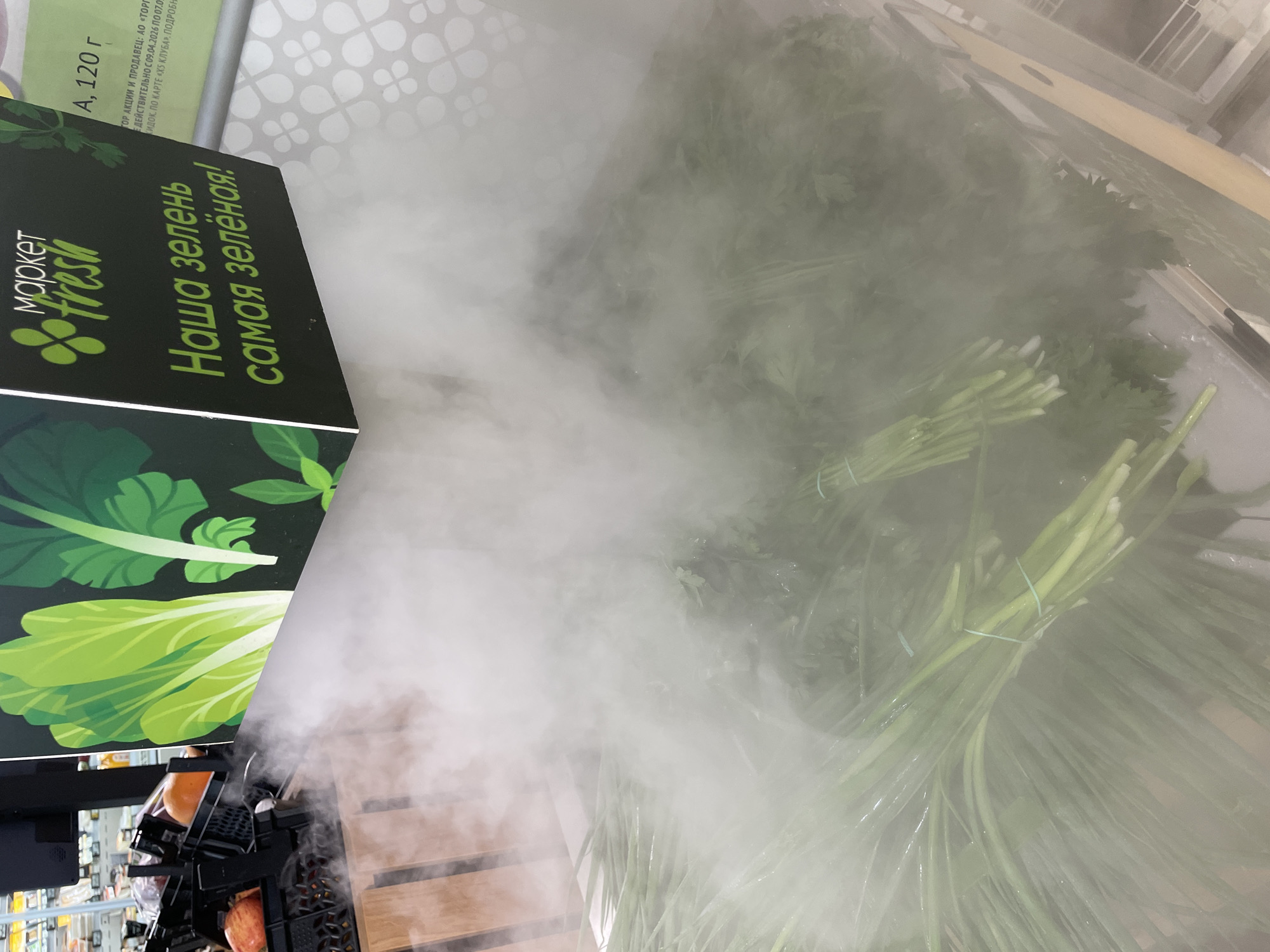 The image shows a display of fresh herbs, likely in a grocery store in Sochi, Russia. The herbs, which appear to be green onions and parsley, are bundled and arranged on a shelf, with a mist of water being sprayed over them to keep them fresh. A sign above the display reads "market fresh" in Russian and features an image of green vegetables. The overall setting suggests a retail environment focused on fresh produce.