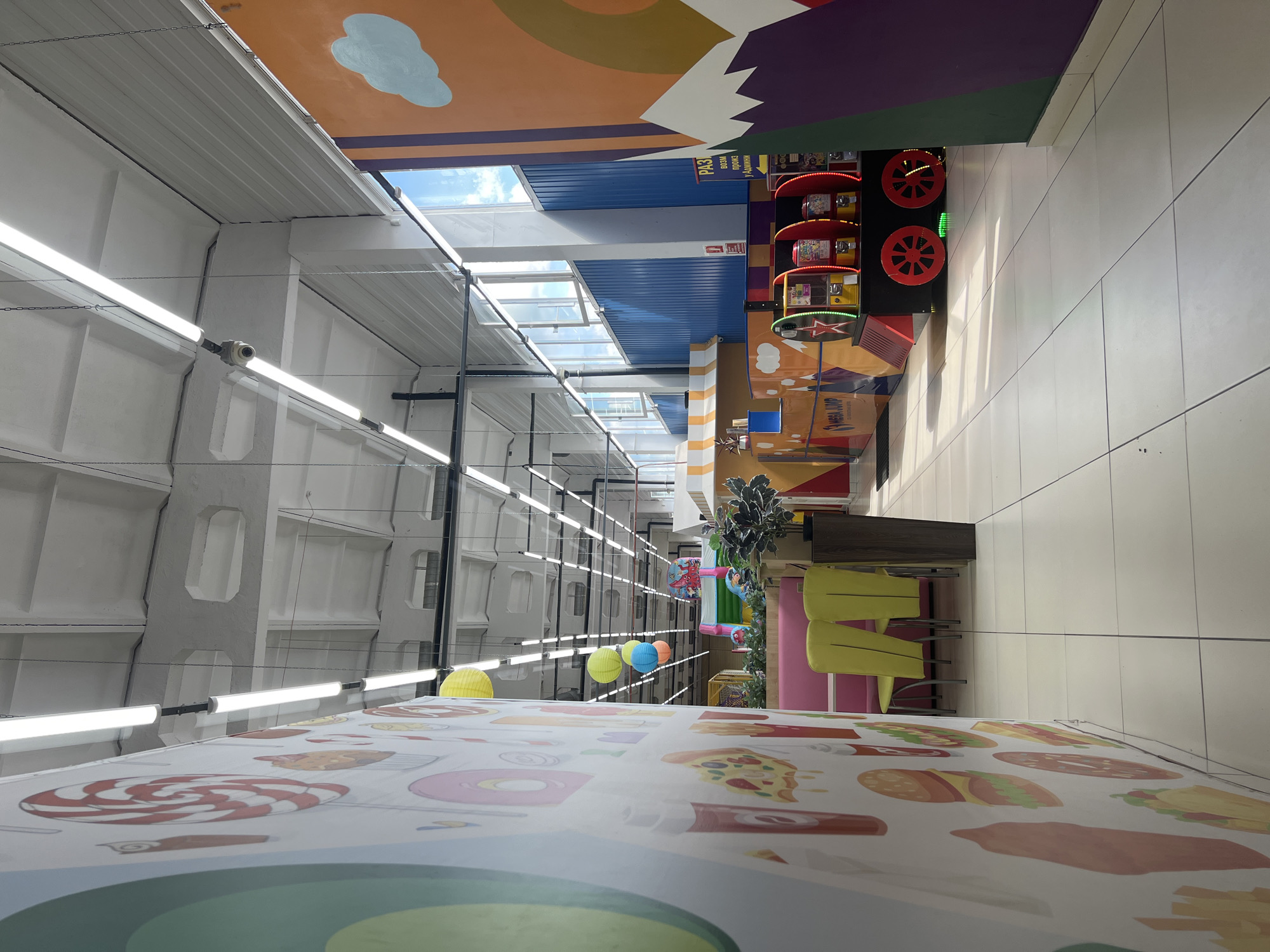 The image depicts the interior of an entertainment center or arcade, possibly located in Chaikovsky, Russia, judging by the presence of Russian text on a sign. The environment is brightly lit by fluorescent ceiling lights and natural light streaming through large windows along the far wall. The foreground is dominated by a wall decorated with large, colorful cartoon images of food items like lollipops, burgers, and pizza, suggestive of a family-friendly establishment.
In the mid-ground, there are seating areas with bright pink booth-style benches and yellow chairs. Beyond the seating, there is a vibrant play area, partially visible, with colorful inflatable structures, possibly a bouncy castle. Several gumball or toy vending machines are lined up, resembling a small train. One machine has text that includes "MEGA JUMP". The floor is tiled with large, light-colored square tiles.
In the background, the ceiling is high and industrial, with exposed beams and conduits, typical of a large public space. The overall impression is one of a lively and stimulating environment designed for entertainment and recreation, particularly for children. There are no people clearly visible in the image.