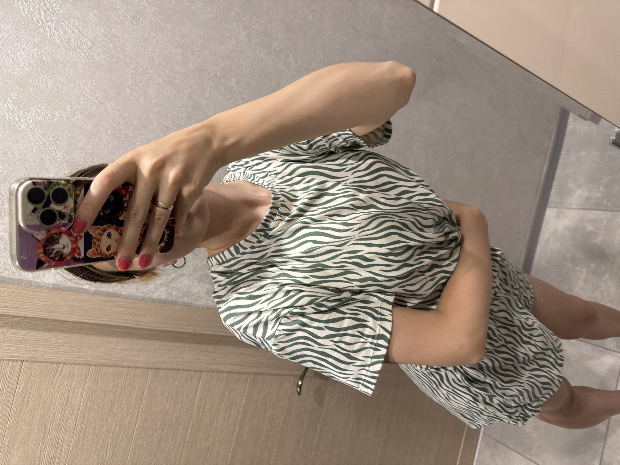 A person stands in front of a mirror, taking a selfie. They are wearing a short-sleeved dress with a green and white zebra print. The dress has a round neckline and appears to be made of a lightweight fabric. The person's right arm is raised, holding a smartphone with a colorful case depicting cats. Their left arm is bent and rests against their stomach. The background is a plain, textured wall and a wooden door with a silver handle. The flooring is tiled. The scene is indoors, likely in a residential setting such as a bathroom or hallway. There is no information to suggest a specific location in Moscow, Russia.