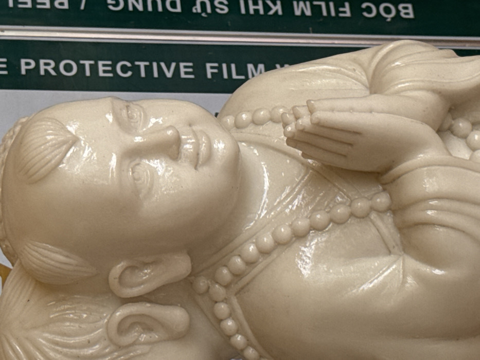 The image displays a detailed close-up of an off-white, lustrous figurine, depicting a juvenile figure. The figure possesses a rounded face with a subtle smile, downcast eyes, and short, stylized hair. Its ears are distinctly sculpted. Draped around its neck and torso is a strand of spherical beads. The figure's hands are pressed together in a prayer-like posture at chest height, above flowing, sculpted drapery. A second, similar figure is partially visible behind the primary subject, with its ear and a portion of its head showing. The background to the right consists of a vertical dark green surface bearing white text that reads "PROTECTIVE FIL" and "BÓC FILM KHI SỬ DỤNG / PEEL". This scene is located in Đăk Hring, Vietnam.