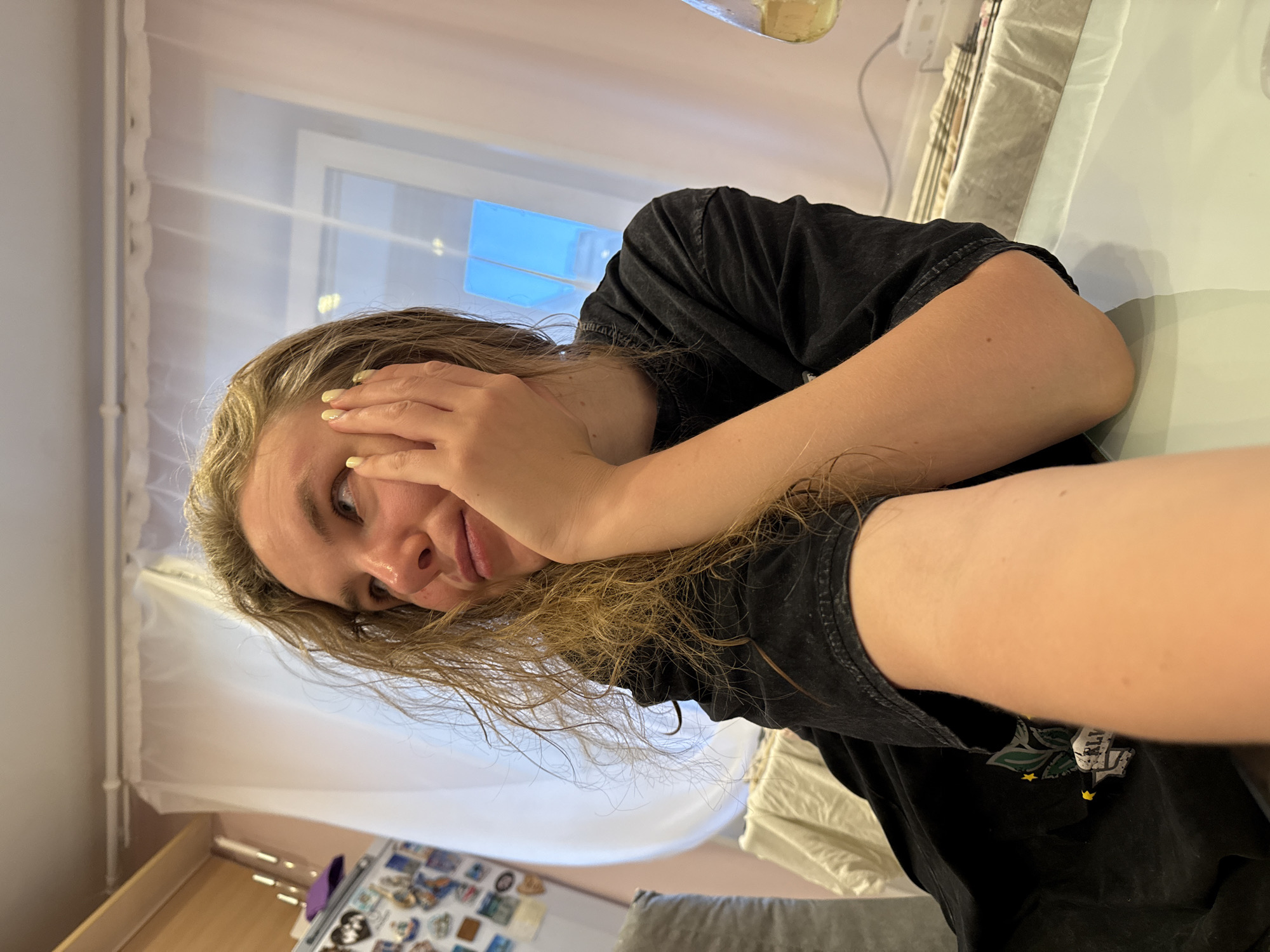 The scene is set indoors, in the city of Vladimir, Russia. A young woman with long, wavy, light brown or blonde hair is positioned in the foreground, facing slightly left of center and looking upwards and to the right. Her skin tone is fair. She is wearing a dark, possibly black or charcoal gray, short-sleeved t-shirt with a relaxed fit. A partial design, featuring green elements and yellow stars, is visible on the left side of her shirt. Her right hand is propping up her chin and lower cheek, with fingers (thumb, index, and middle) lightly touching her face. Her fingernails are painted with a pale yellow polish. Her right elbow rests on a glossy, light-colored table surface, which reflects ambient light. Her left arm extends towards the bottom of the frame, partially out of focus.
Behind the woman, a window is visible, covered by a sheer white curtain. Through the curtain, a bright blue reflection, possibly from the sky, is visible through the windowpanes. To the left side of the frame, behind the woman, a white refrigerator is covered with numerous colorful magnets. Above the refrigerator, a light-colored wooden shelf or cabinet is mounted, holding several thin, vertical objects resembling candles in holders, along with a small purple item. To the far right, a portion of a glass object, possibly a beaker or bottle containing a yellowish liquid, is visible on the table. A light-colored, patterned fabric, possibly a tablecloth or runner, is draped over a surface behind the table. The lighting is bright and natural, emanating from the window.