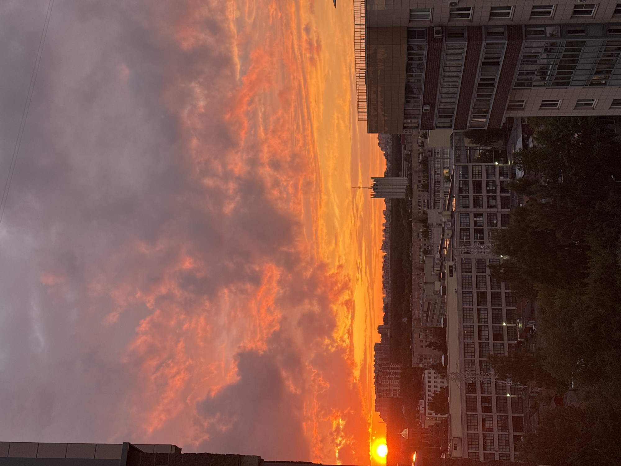 A wide shot captures a vibrant sunset over an urban landscape in Unknown, Russia. The sky is filled with dramatic clouds painted in fiery hues of orange, red, and pink, transitioning to a softer purple at the top. The sun, a bright orange orb, is visible on the left side of the frame, low on the horizon, casting a warm glow over the scene.
Below the striking sky, the cityscape unfolds. In the foreground on the right, a multi-story building with numerous windows and a balcony area dominates. To its left, a large, industrial-looking building with many windows stretches across the mid-ground. Beyond this, more buildings of varying heights form a dense urban fabric. A distinct, tall, monolithic tower with a pointed top stands out in the distance, towards the center of the image.
The lower part of the image shows the tops of trees, indicating greenery within the city. The overall impression is one of a city bathed in the dramatic light of a sunset. There are no people or visible animals in the frame. The scene is static and serene.