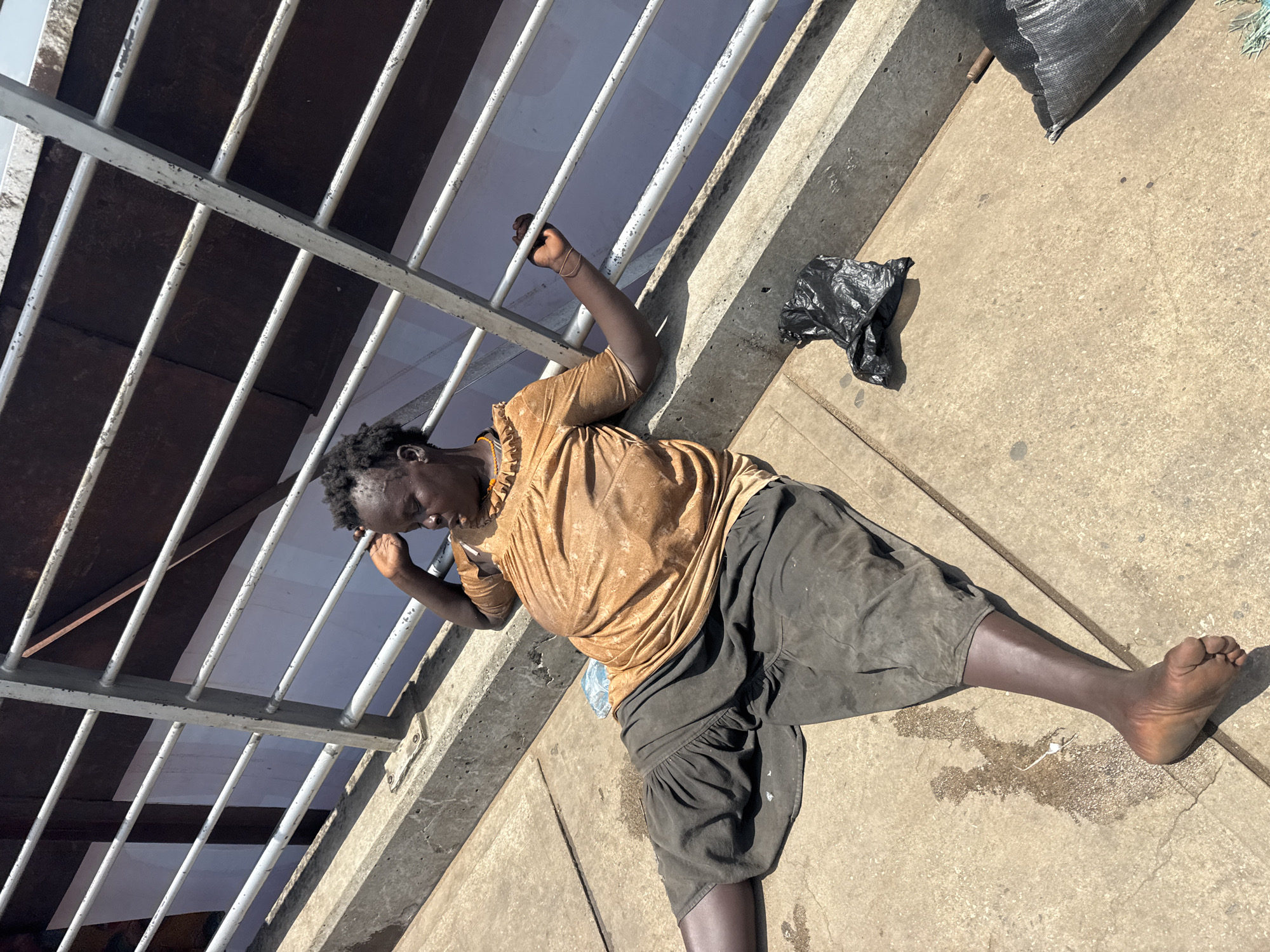 The image, taken with a tilted perspective, captures a person lying on a concrete surface in bright daylight. The subject is an individual with dark skin and short dark hair, wearing a light brown or gold short-sleeved shirt and dark grey shorts, both appearing worn. They are barefoot. The person lies on their left side, curled slightly, with their right arm extended and hand gripping a vertical bar of a metal railing. Their left hand is near their head. A clear plastic water bottle is partially visible underneath their torso.
The concrete ground around the person shows some stains and a wet patch near their bare feet, with a thin metal rod or pipe also visible there. A crumpled black plastic bag rests near the person's head, and another, larger black bag or sack is seen further back on the right side of the frame.
Above and beside the person runs a metal railing structure, consisting of vertical bars held by a horizontal beam. These bars are light grey or white, with visible wear and peeling paint. Behind the railing, there's a darker area, possibly a ceiling or roof, and a lighter area beyond, suggesting an open space.
The strong shadows indicate a sunny outdoor environment, likely mid-day. No text is visible in the image. The scene depicts an individual resting or incapacitated in a public, urban setting, consistent with the given location context of Onitsha, Nigeria.