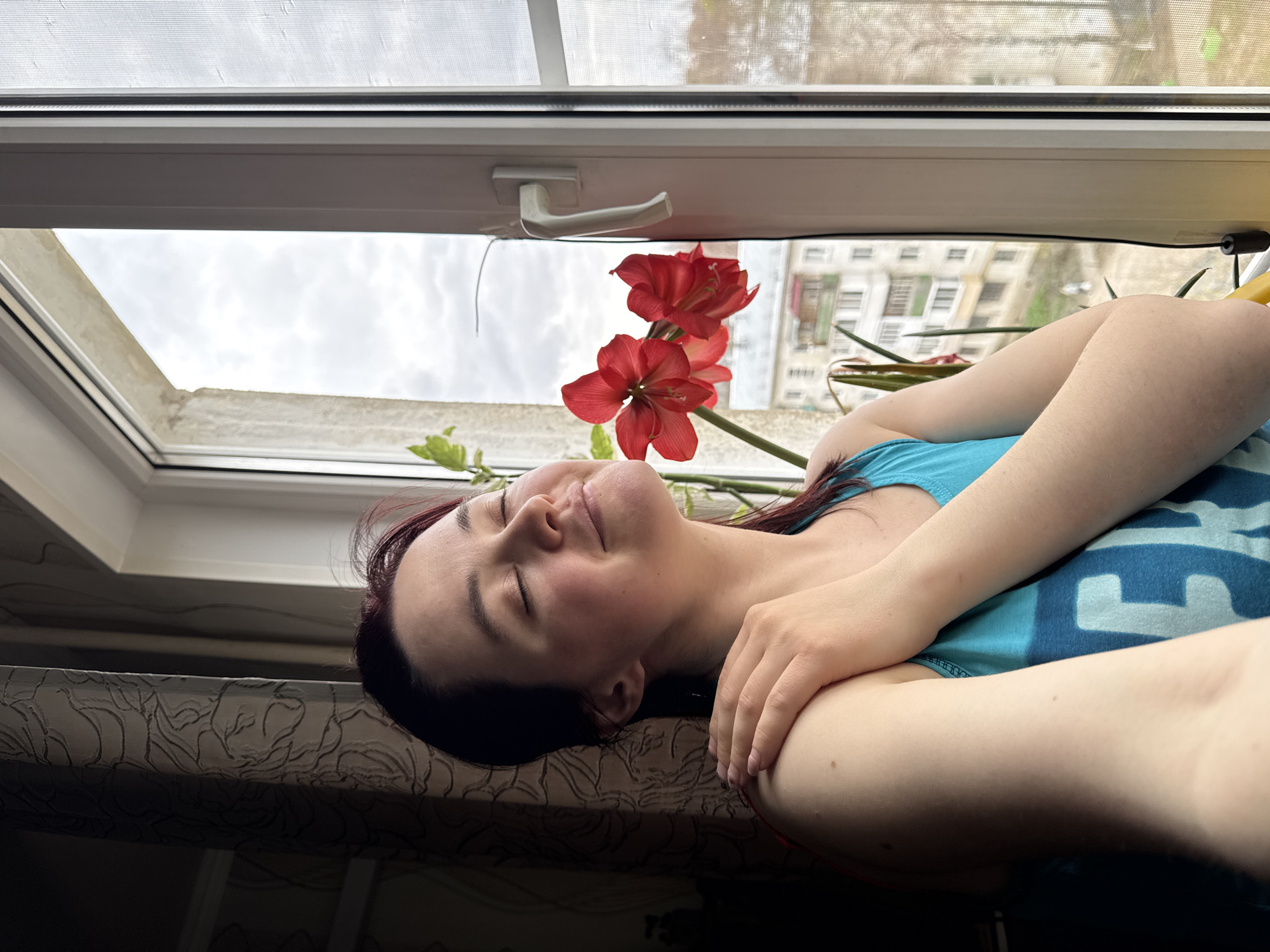 An indoor scene in Izhevsk, Russia, depicts a person resting by a window on a cloudy day. The image is taken from a slightly elevated, rotated perspective, showing a person, likely female, with dark red hair, lying on their back with closed eyes, suggesting sleep or deep relaxation. Their head is positioned towards the left of the frame, resting on a patterned, light-colored surface, possibly a windowsill or bench. They wear a light blue tank top with a partial, indistinct dark blue graphic or text on the chest.
Adjacent to the person's head, two vibrant red flowers with visible stamens, likely amaryllis, are blooming in a pot on the windowsill. Green foliage from the plant is also visible. The large window above the person has a white frame and a visible white handle. The upper section of the window includes a mesh screen.
Through the window, an urban landscape is visible under an overcast sky filled with white and grey clouds, indicating a cloudy day, possibly late morning or afternoon due to the diffused light. The background features multi-story residential buildings, characteristic of city architecture. The window frame and the outer sill show some signs of dust or dirt. The overall mood conveyed is one of peacefulness and rest amidst an urban setting. No legible text is visible on the person's clothing or elsewhere.