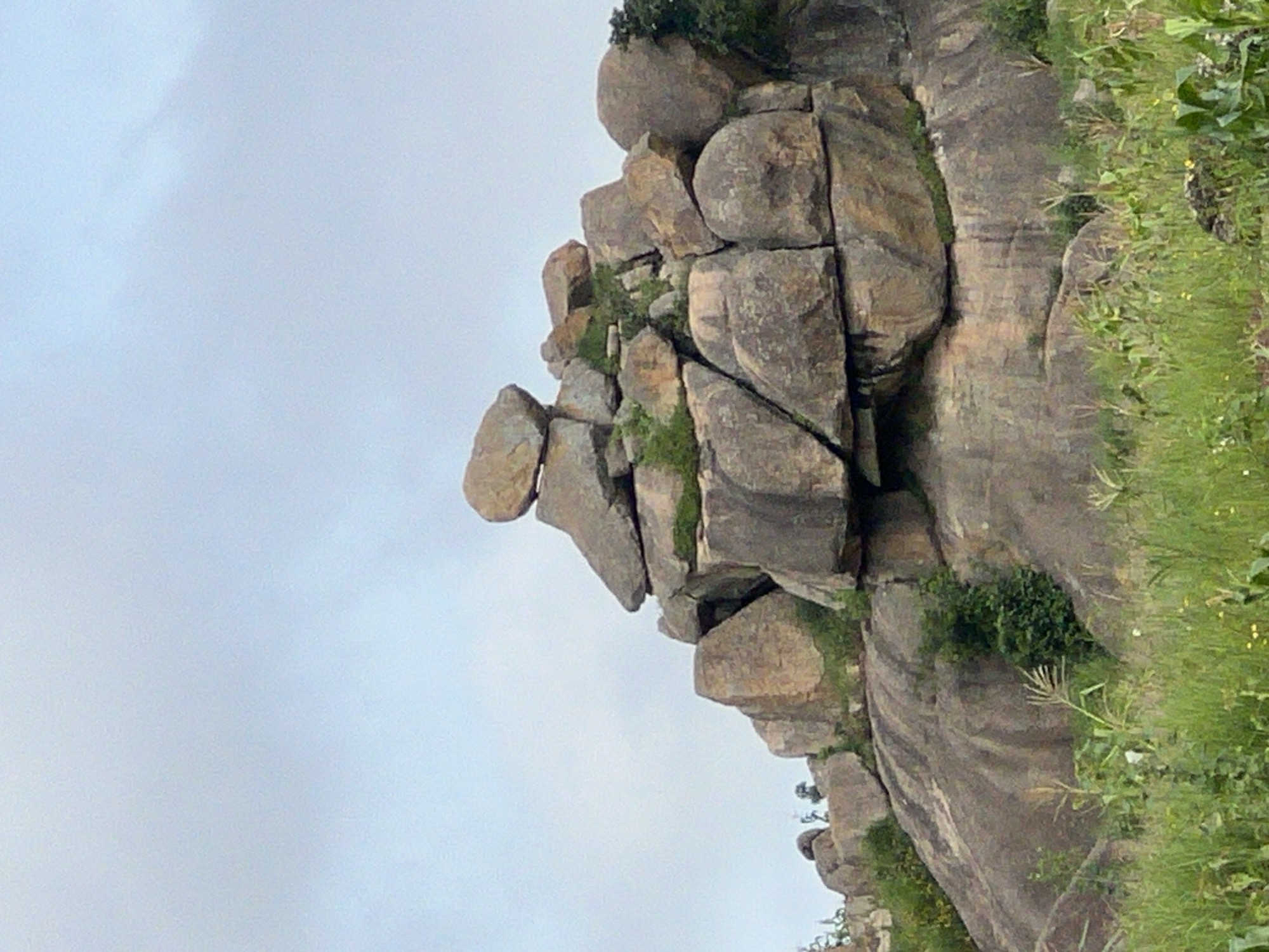 This image depicts a large rock formation situated on a grassy incline, under a pale, overcast sky. The rock formation is composed of numerous large, weathered boulders, some of which appear precariously balanced on top of one another. Patches of green vegetation, likely moss or small shrubs, cling to the crevices and surfaces of the rocks. The foreground is filled with tall, green grass, some of which has dried out, interspersed with sparse green foliage. The overall scene suggests a natural, undeveloped landscape. This formation is likely located in the city of Jos, Nigeria, a region known for its rocky terrain and granite outcrops.