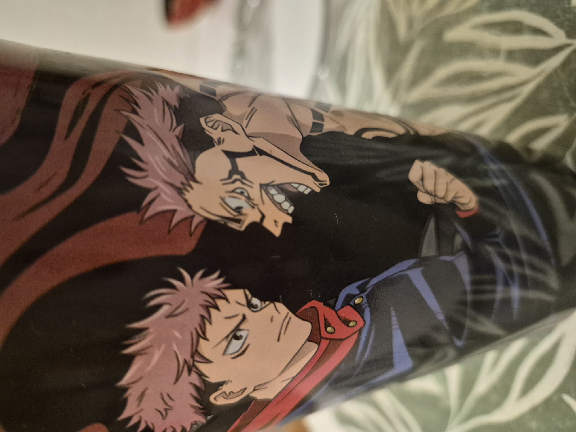This is a close-up, angled, full-body photograph of a cylindrical object, likely a cup or bottle, decorated with an anime-style illustration. The illustration features two male characters with spiky pink hair, facing each other. The character on the left has a neutral expression, wearing a blue jacket with red trim and gold buttons. The character on the right has a more aggressive expression, with visible teeth and markings on their face and body. The background of the illustration is black with red, wavy lines. The object is placed on a surface covered with a green and white patterned fabric. The lighting suggests an indoor setting, and there is no indication of a specific city or location.