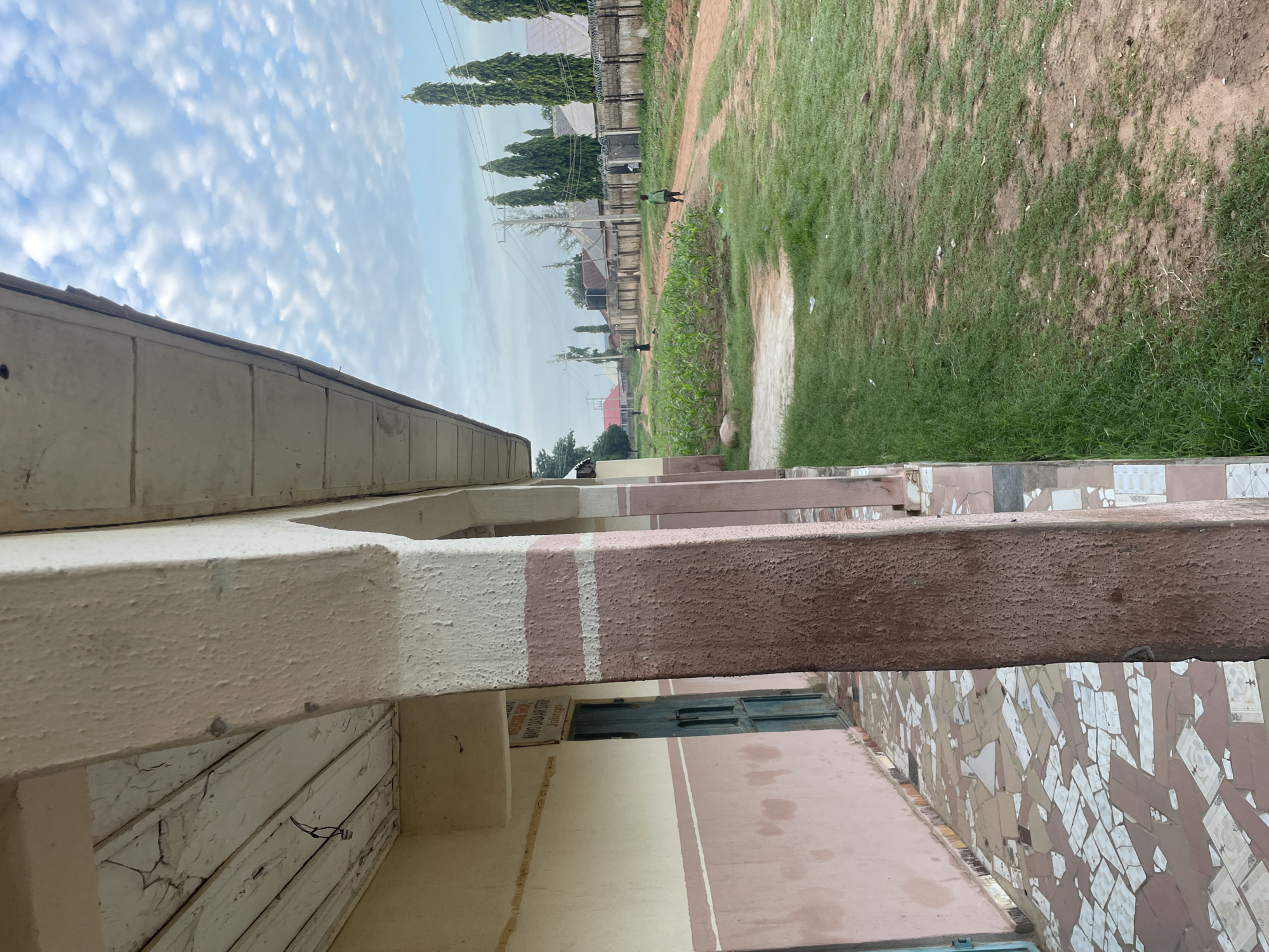 The image shows a dusty road leading through a residential area in Gbaza, Nigeria. The road is lined with a small cornfield on the left and a grassy area on the right. In the background, houses with corrugated iron roofs are visible, as well as a concrete wall with barbed wire on top. Two young children, one in a green shirt and the other in a dark outfit, are walking along the road. The scene is illuminated by the soft light of a cloudy sky. The foreground features a covered walkway with columns, a tiled floor, and a pink and cream colored building. A blue door is visible on the left side of the walkway, and a sign above it reads "MAYO GASA AIR STRIP, JALINGO". The overall impression is one of a simple, rural environment.