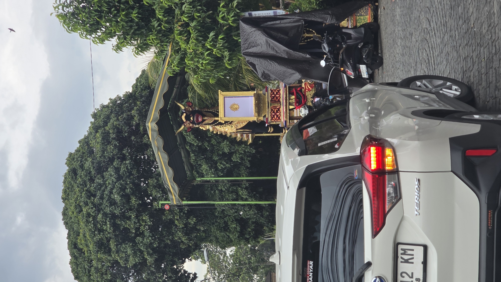 This image captures a street scene, likely in Ubud District, Indonesia, featuring elements of a traditional Balinese Hindu cremation ceremony. In the foreground, the rear of a white SUV is prominently displayed. It is identified by the "TERIOS" emblem on the tailgate, and stickers reading "ASTRA DAIHATSU HANYAR" and "PROTERA Protection." A partial view of its license plate shows "12 KM," and its right taillight is illuminated.
Behind the car stands an elaborate, golden ceremonial structure, characteristic of a Balinese "Bade" or "Ngaben" tower used in cremation ceremonies. This ornate float features intricate carvings, including what appears to be a mythical creature or deity's head at the front, possibly a Nage or Garuda. A large, gilded rectangular platform, potentially a sarcophagus or a "Lembu" (ox effigy) container, sits atop the main structure. The entire setup is positioned under a dark green canopy with yellow trim, supported by green poles.
To the right of the ceremonial float, a black motorbike is partially visible, covered by a black tarp. The background is dominated by dense, lush green trees, suggesting a tropical environment. The sky is overcast and cloudy, indicating a muted light condition during the daytime. A small bird can be seen flying in the upper left corner. The ground appears to be paved, possibly with patterned stones. The overall scene depicts the preparation or a segment of a significant cultural and religious event, specifically a Balinese Ngaben ceremony.