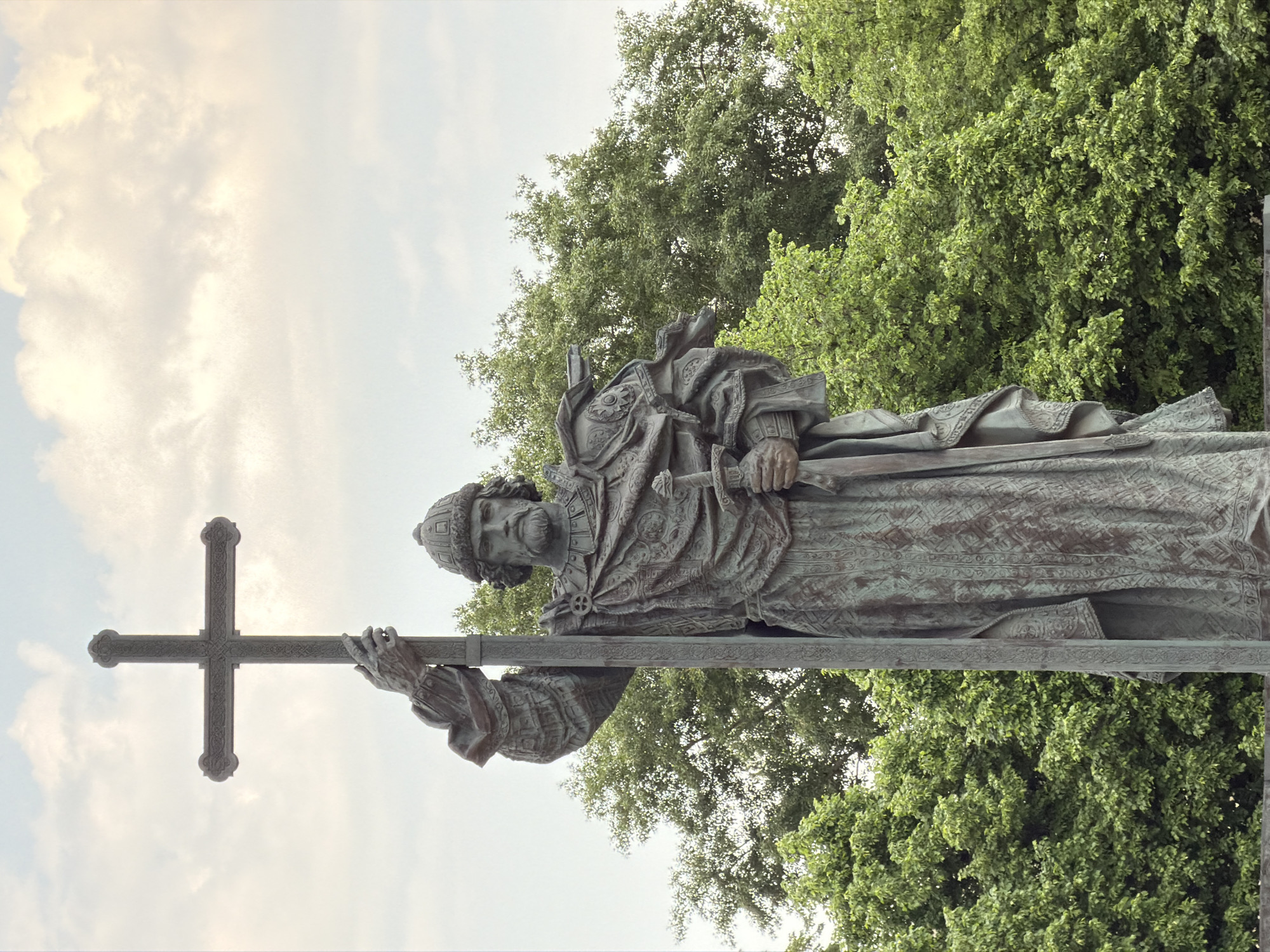 The image shows a bronze statue of Patriarch Hermogenes holding a large cross. The statue is located in Moscow, Russia. It depicts a man in ornate robes and a hat, gripping the cross with both hands. The background consists of green trees and a cloudy sky.