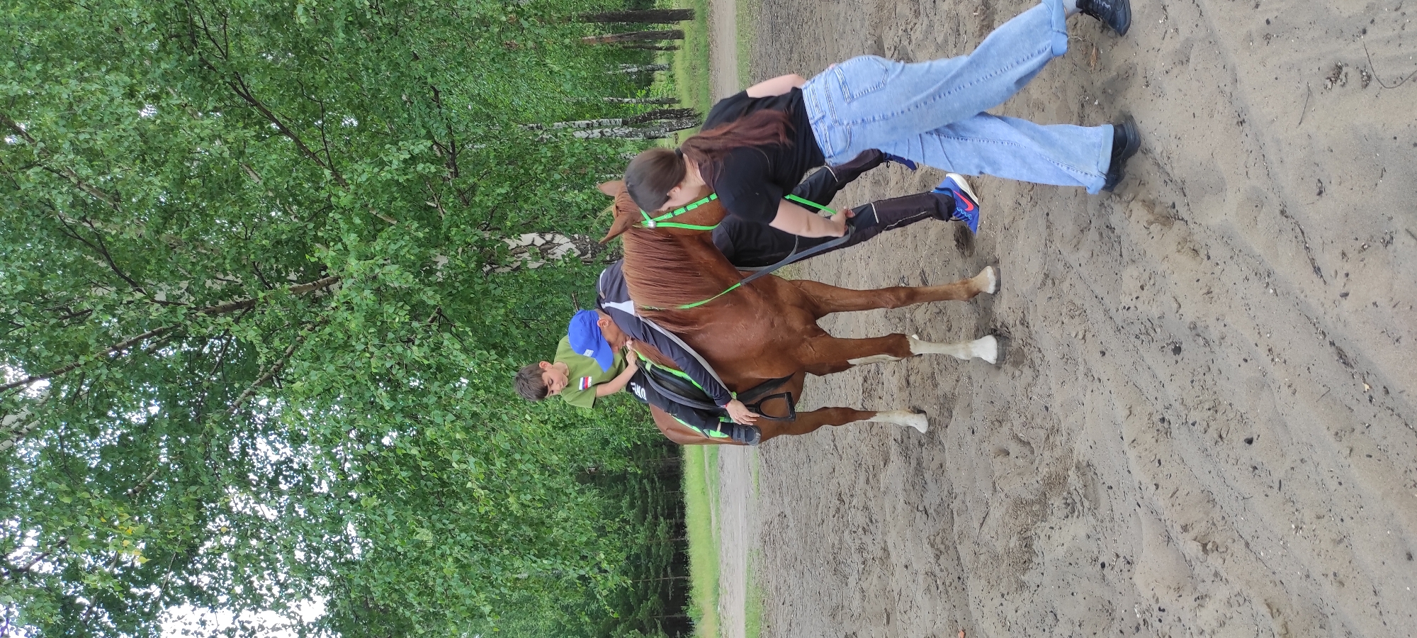 In an outdoor setting, likely Noviy, Russia, with trees and sandy ground visible, two people are interacting with a brown horse. The horse is equipped with a saddle and a bright green bridle. A young person wearing a green t-shirt and a blue baseball cap is seated on the horse, holding onto its tail. Another person, dressed in a black t-shirt and light blue jeans, stands beside the horse, holding onto a rope attached to its bridle and appears to be guiding or assisting the rider. The weather seems overcast, and the scene suggests a moment of equine instruction or leisure activity. The time of day is not explicitly indicated but the lighting suggests daytime. There is no visible text in the image.